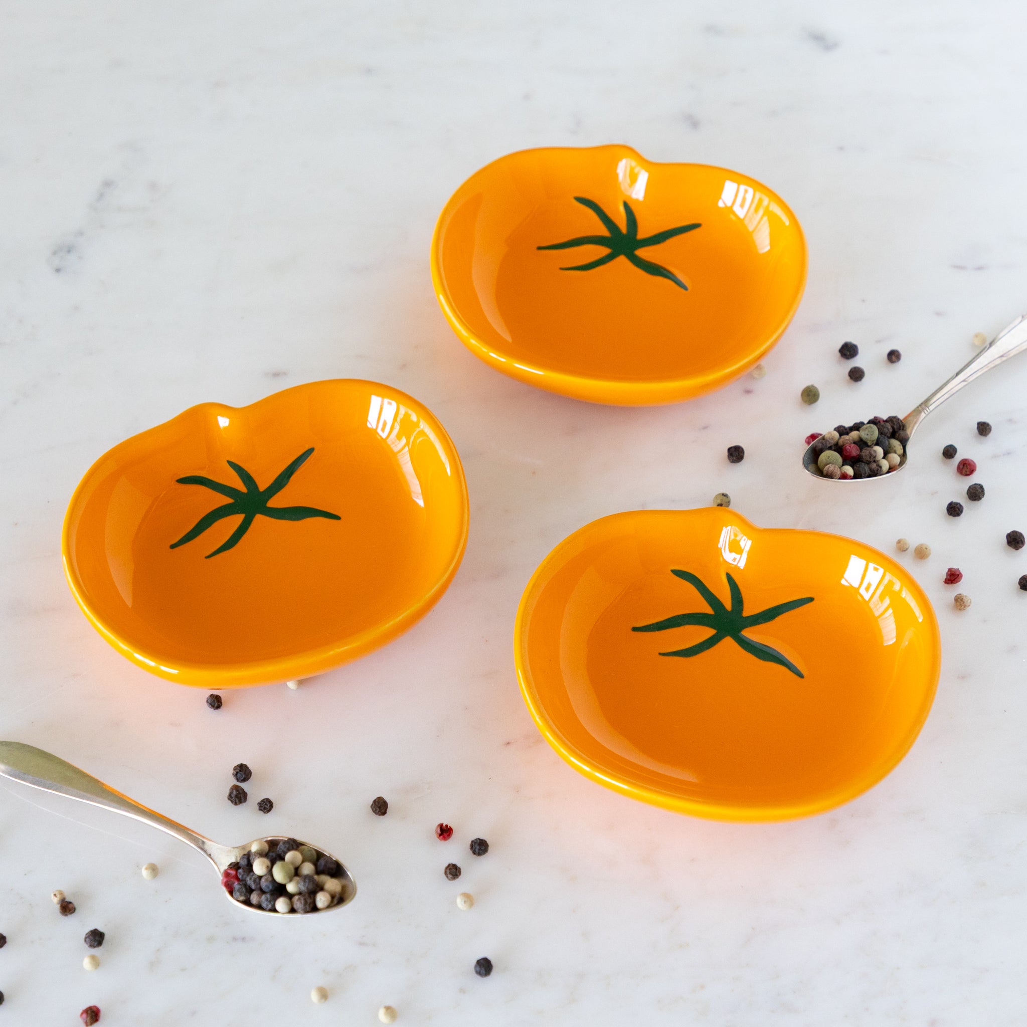 Three orange ceramic dishes shaped like tomatoes on a marble surface with peppercorns.