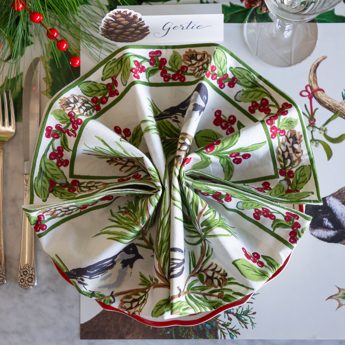 A folded white napkin with a woodland bird and berry print is arranged on a place setting table runner, surrounded by gold-toned flatware, pinecones, evergreen branches, and a glass.