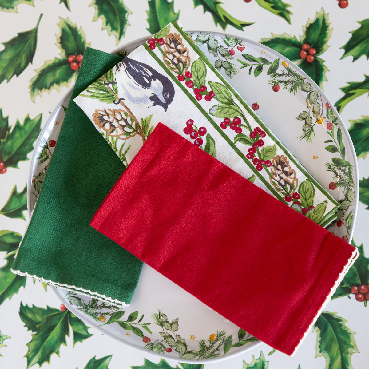 A styled flat-lay of Christmas-themed table linens, including a solid dark green napkin, a red napkin, and a printed dinner plate featuring a bird, pinecones, and holly leaves on a white background with a matching holly pattern.