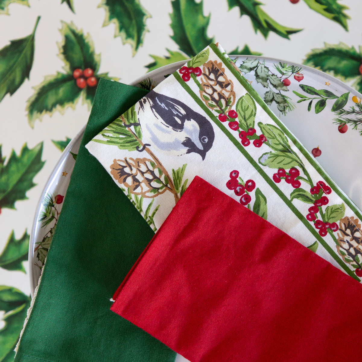 A styled flat-lay of Christmas-themed table linens, including a solid dark green napkin, a red napkin, and a printed dinner plate featuring a bird, pinecones, and holly leaves on a white background with a matching holly pattern.