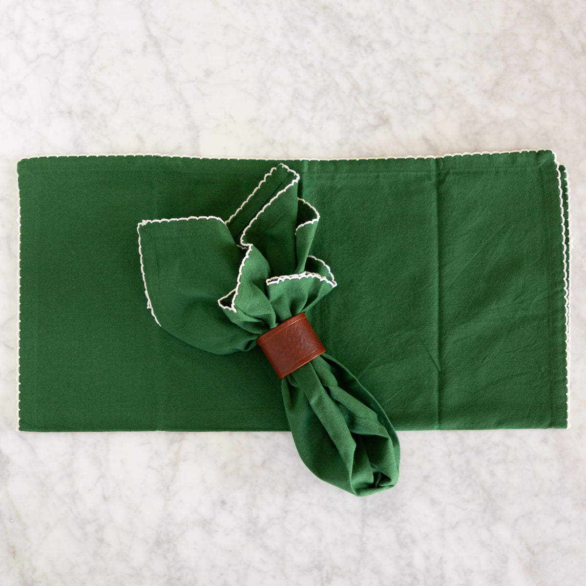 A dark green fabric napkin with white embroidered trim, gathered and secured with a brown leather napkin ring on a light gray marble surface.