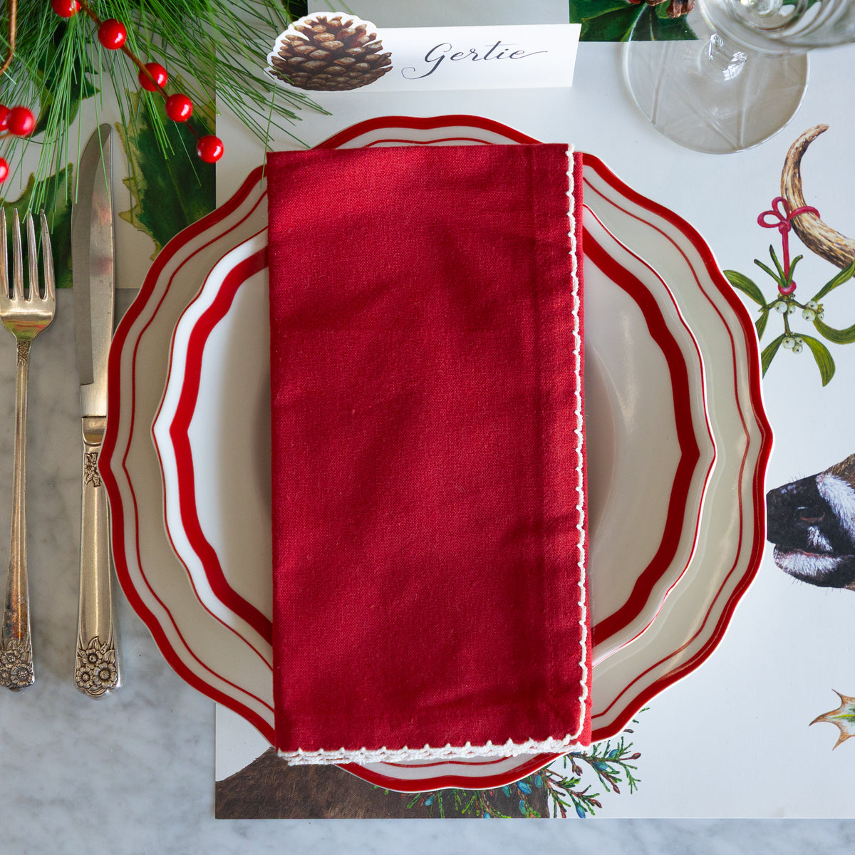 A festive holiday table setting featuring a deep red fabric napkin with white scalloped edges, layered over white plates with red borders, alongside gold-toned cutlery and holiday greenery.