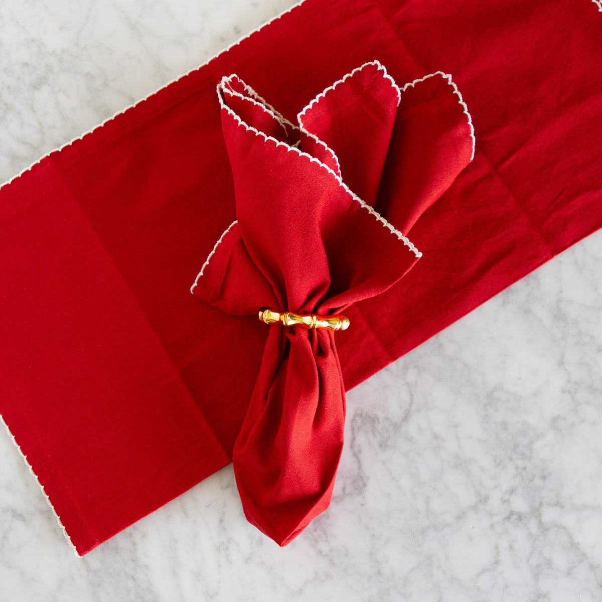 A folded red napkin with white scalloped edges tied with a gold beaded napkin ring, placed on a matching red table runner over a white marble surface.
