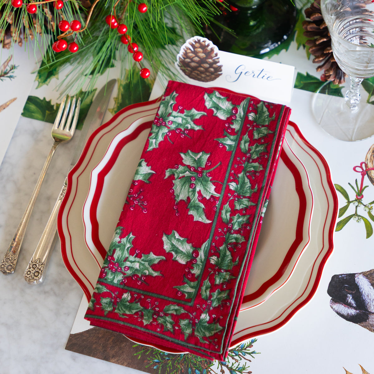 A festive holiday table setting featuring a red napkin with a green holly and berry pattern, layered on a cream and red scalloped charger plate. The setting includes gold-toned flatware, a glass, pinecones, evergreen branches, and a place card reading &