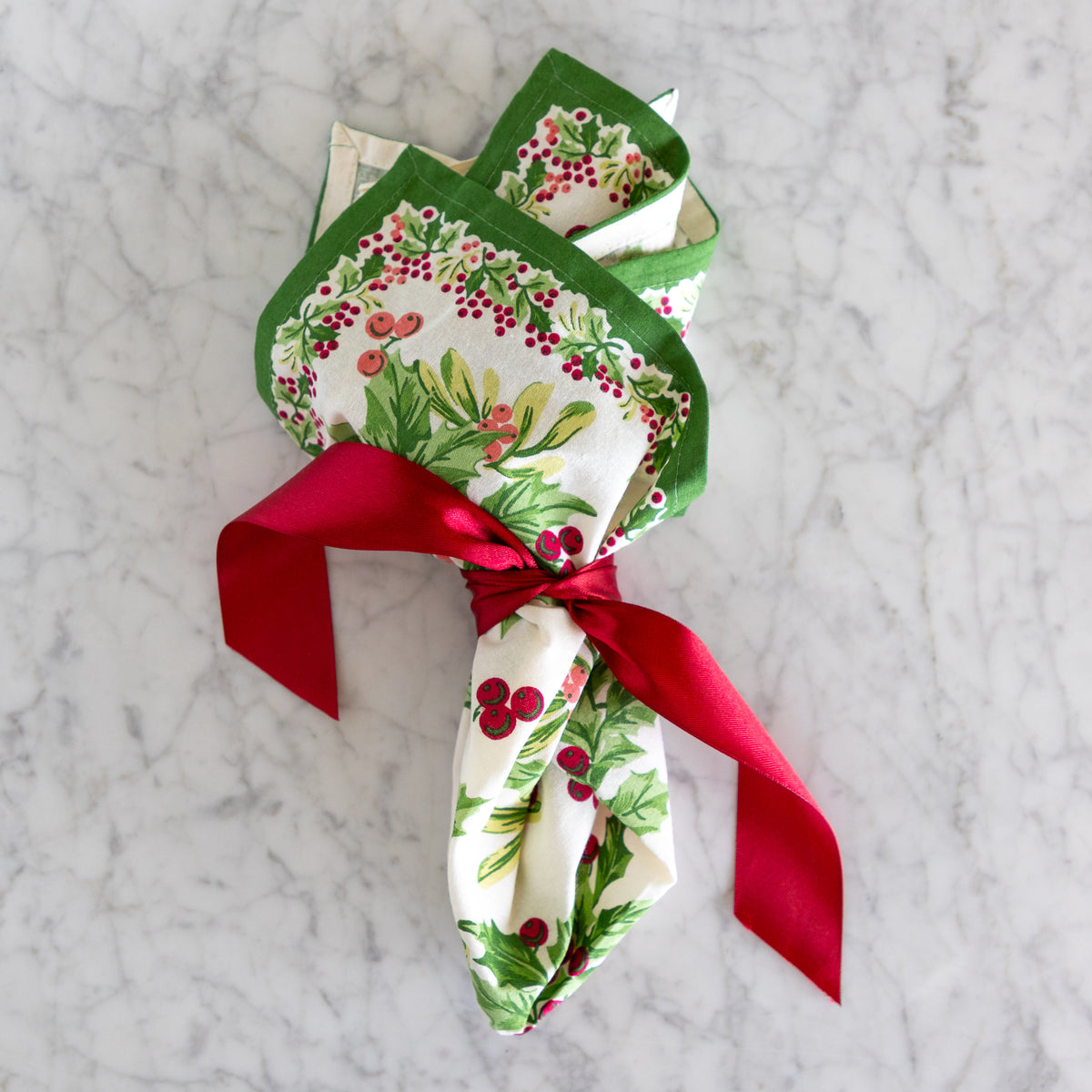 A square Christmas-themed tablecloth with a matching folded napkin and a red ribbon bow, displayed on a light gray marble surface. The fabric features a green border and a pattern of holly leaves and red berries on a white background.