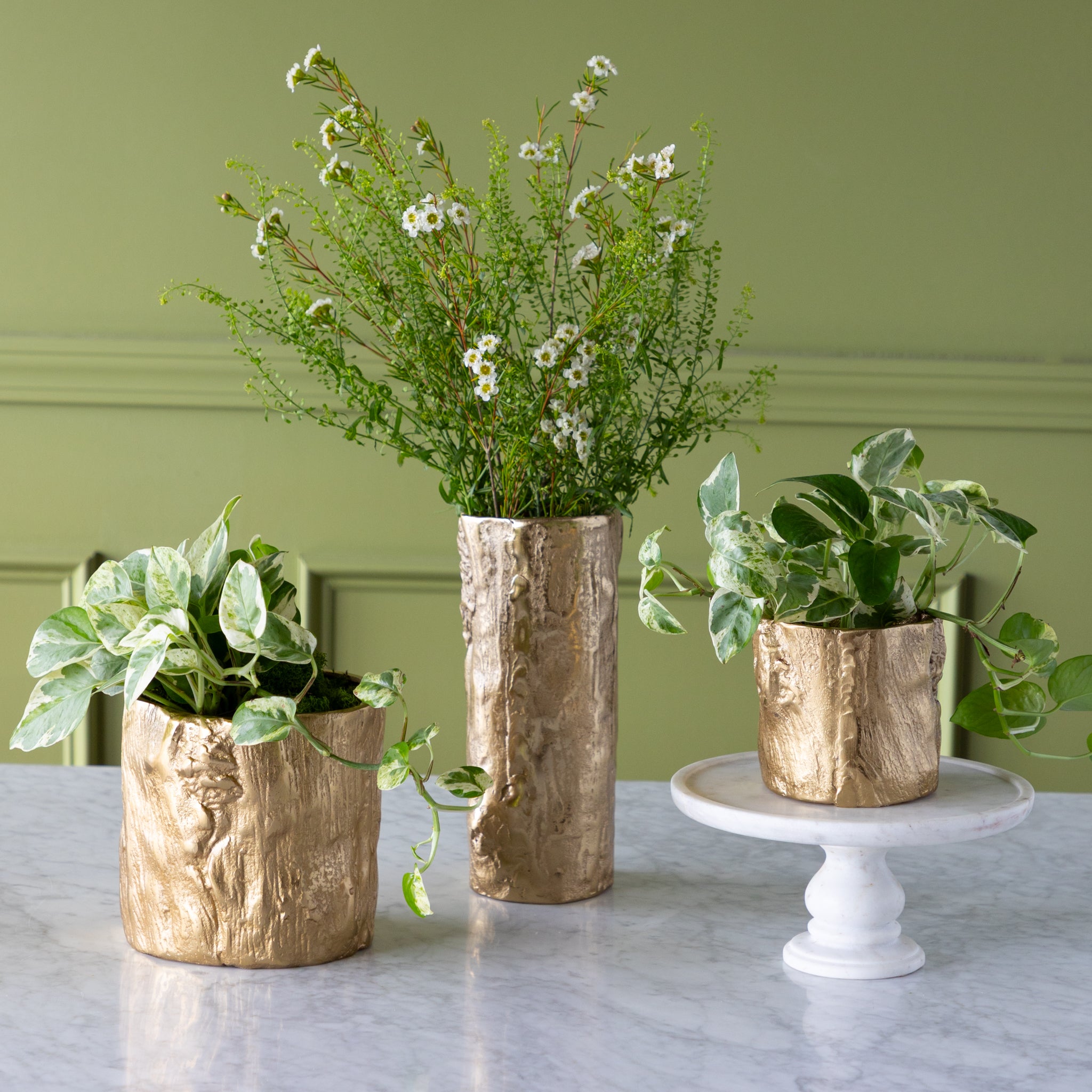 Three Gold Birch Metal Vases with flowers in them on a marble surface with a green wall background.