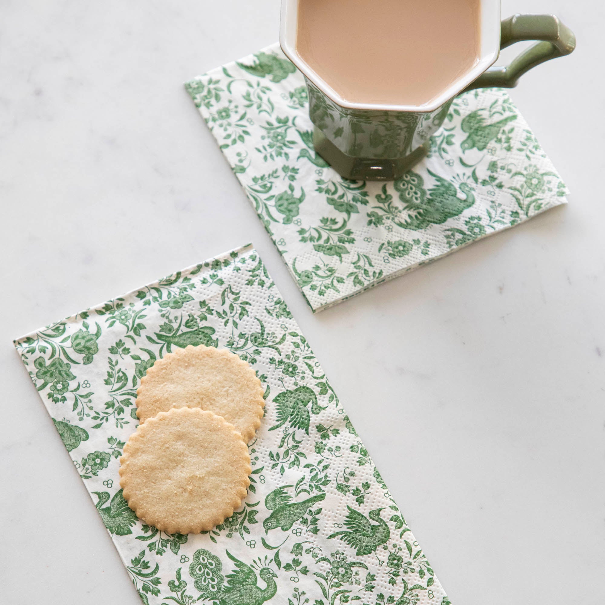 A cup of coffee and cookies on a table adorned with Hester & Cook Green Regal Peacock napkins.