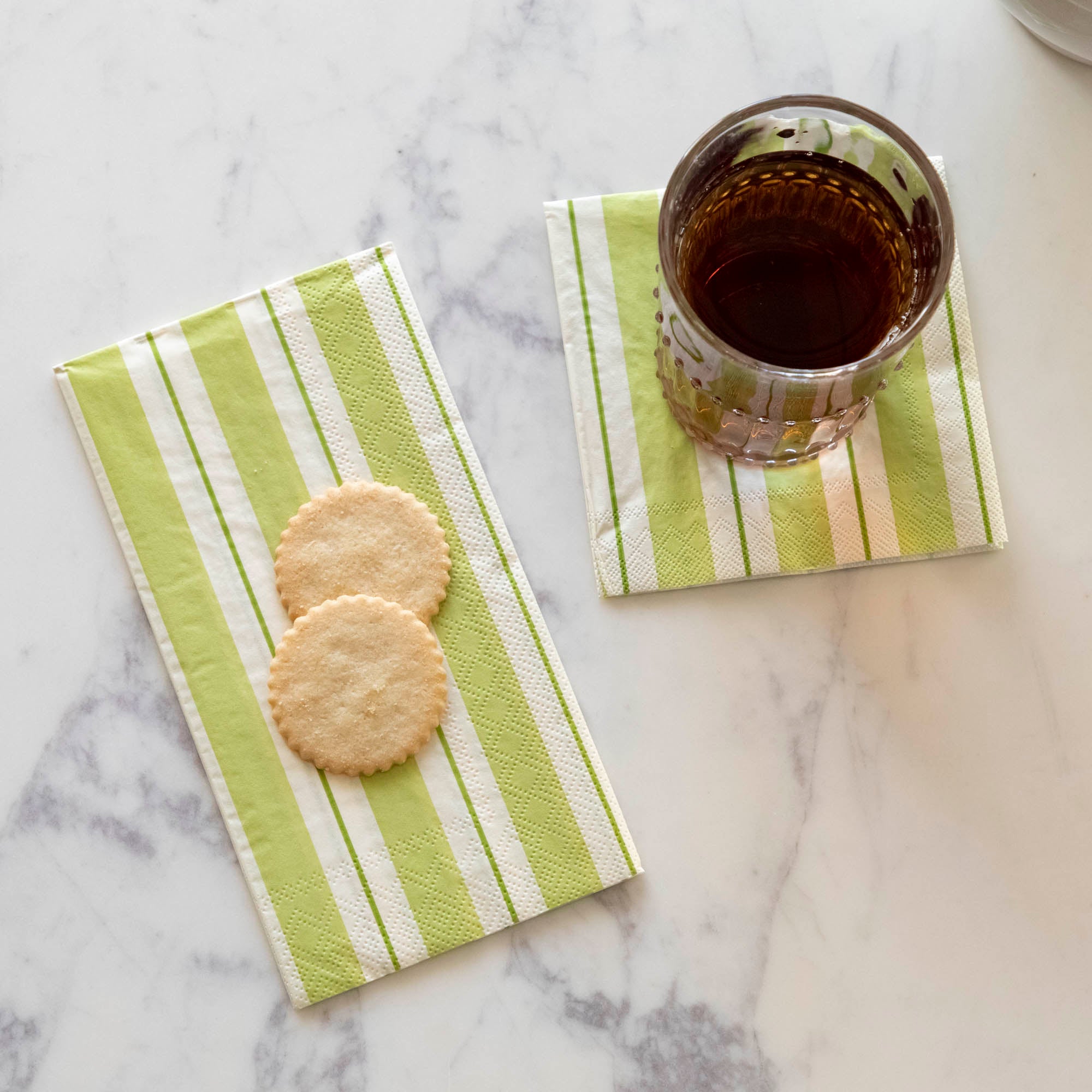 Two Green Awning Strip Napkins on a white table: a Guest napkin with two sugar cookies on it, and a Cocktail napkin under a glass.