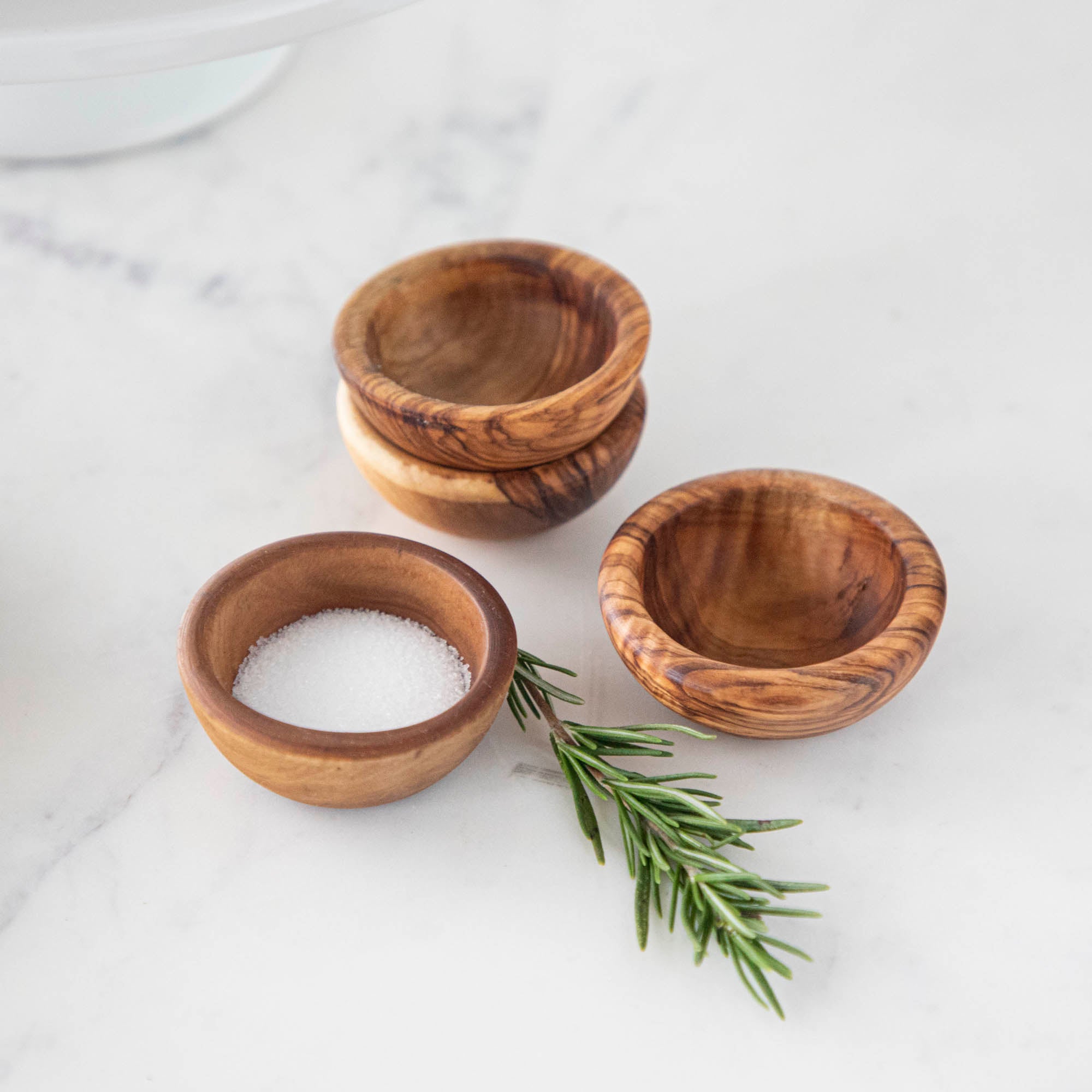 Four Natural Olivewood Pinch Bowls with salt and a sprig of rosemary on a kitchen counter.