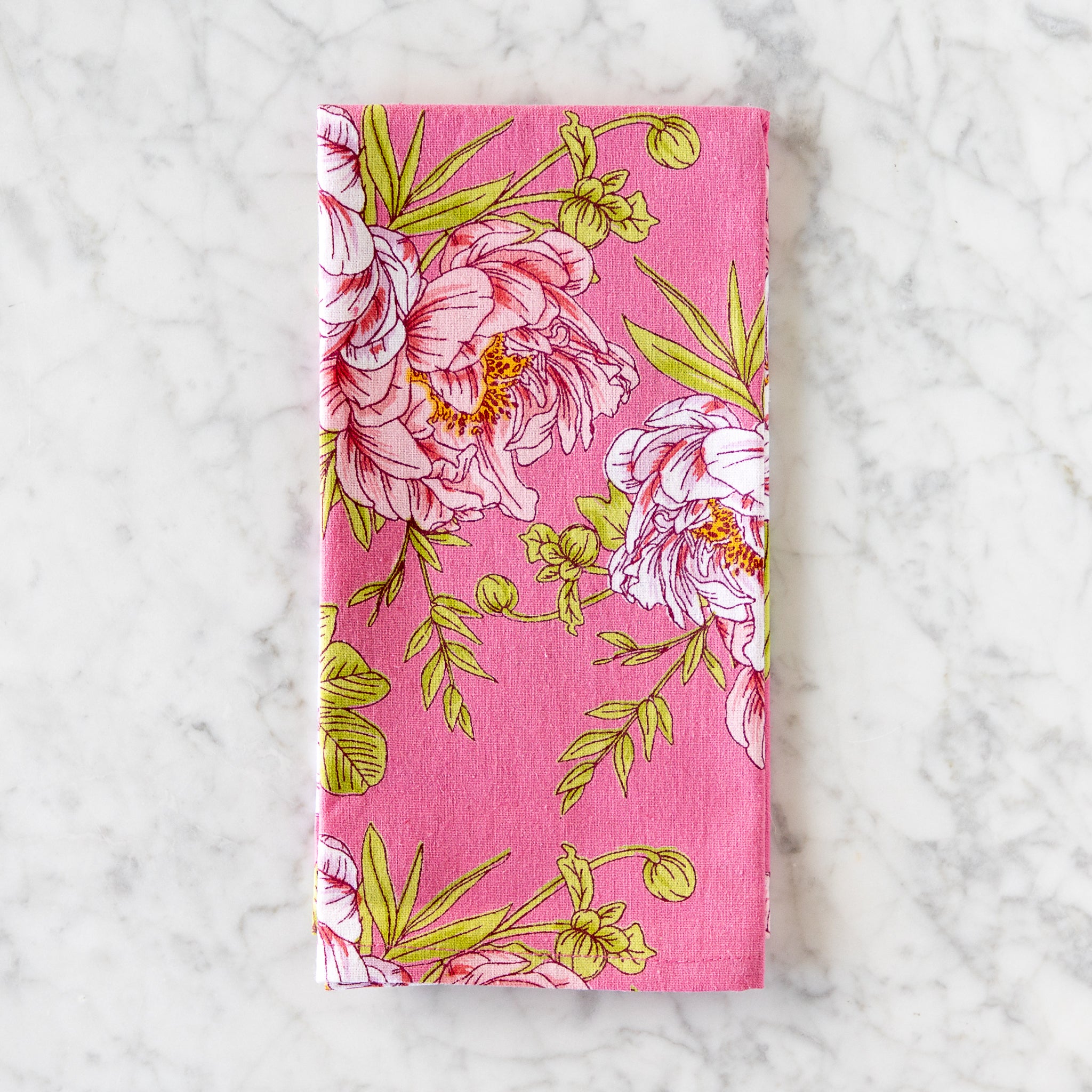 A pink cotton napkin, made in India, features a colorful illustration of pink flowers and green leaves and is displayed on a marble table.