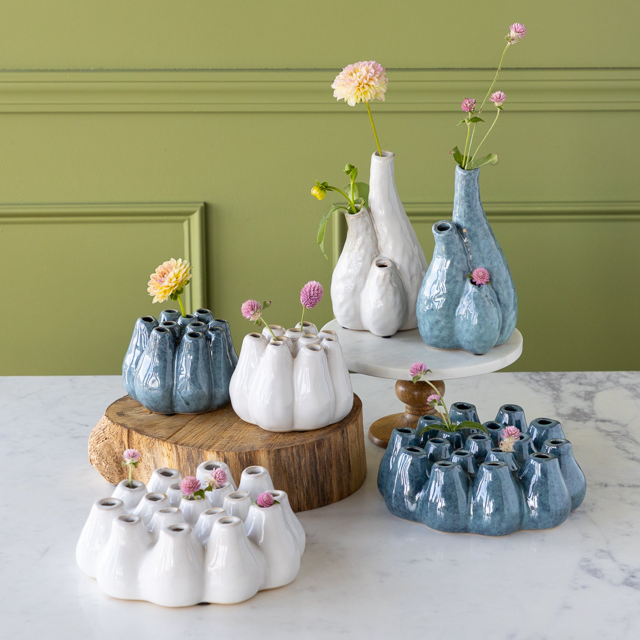 Collection of ceramic vases with flowers on a marble surface against a green wall.