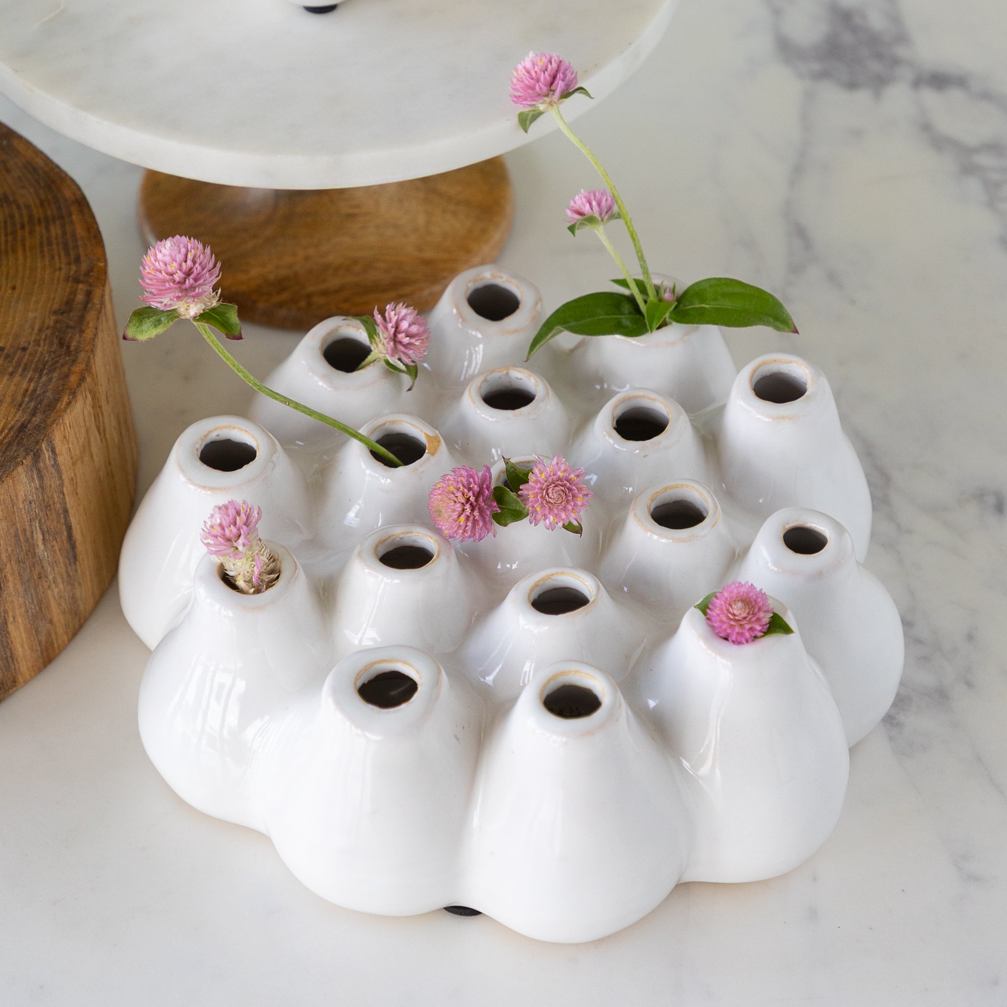 A white ceramic vase with 16 holes and pink flowers on a marble surface.