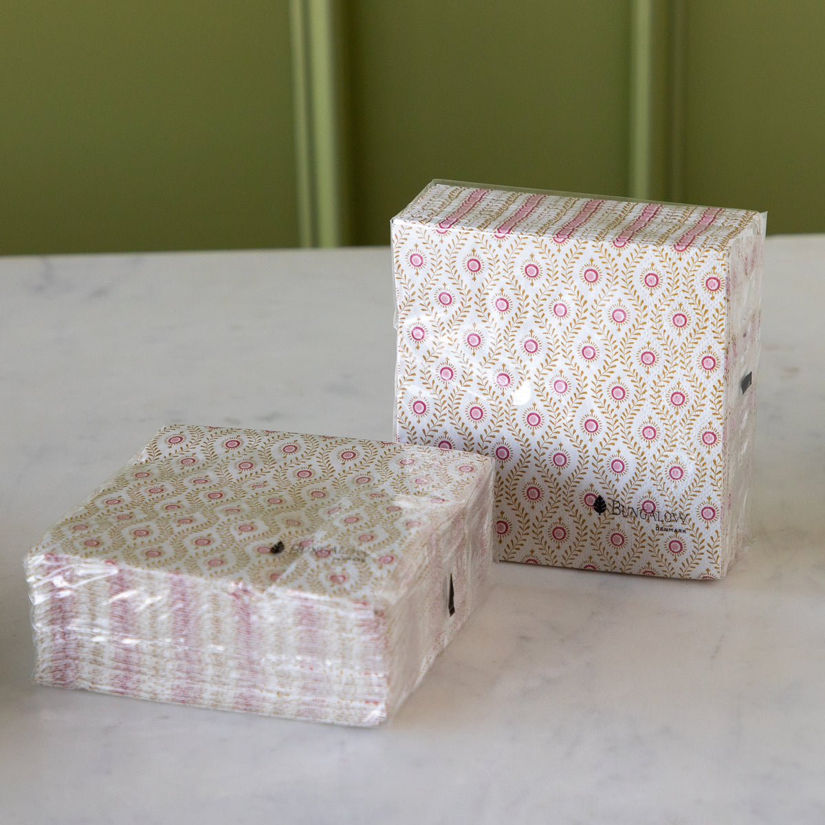 Two stacks of patterned paper napkins wrapped in clear plastic, placed on a white marble surface against a green paneled wall.