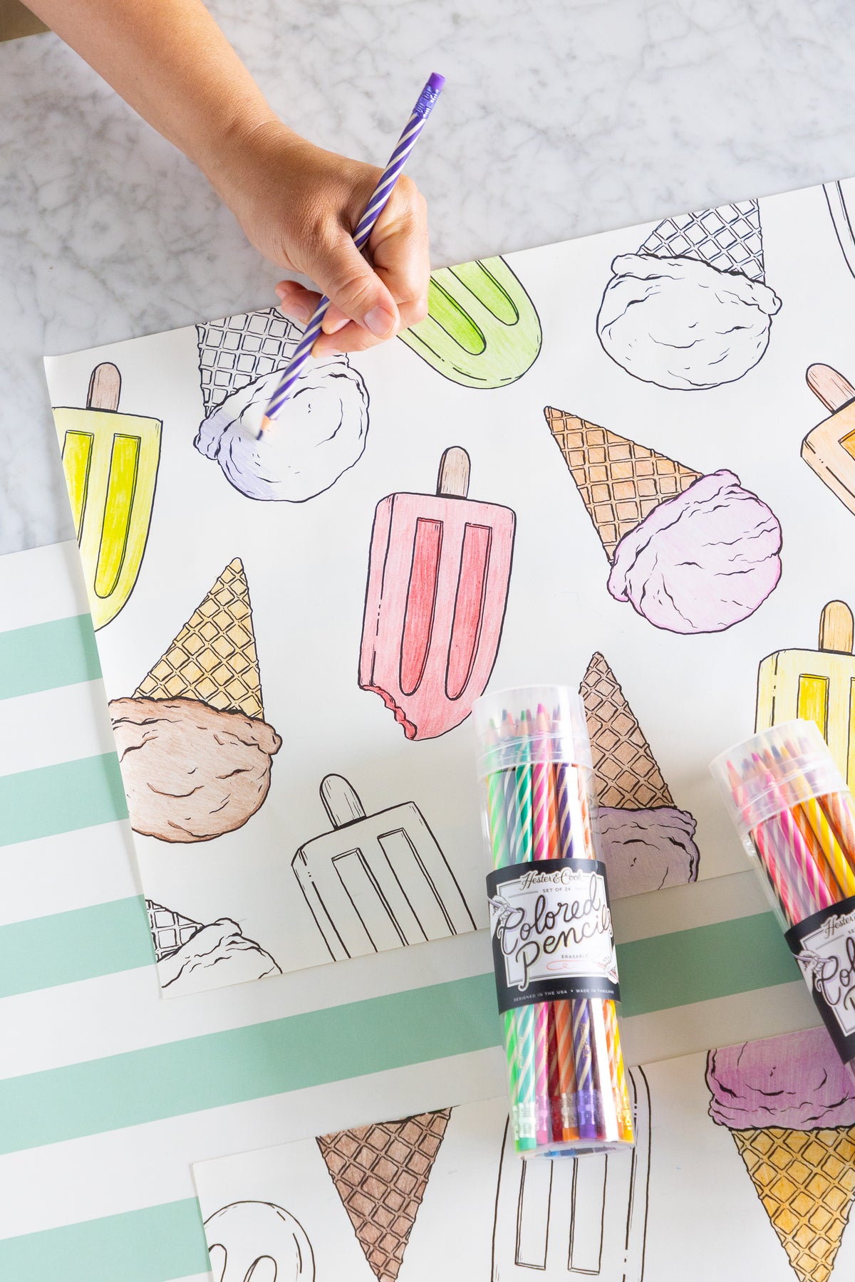 Coloring placemats with ice cream and popsicle illustrations and colored pencils on a striped table runner.