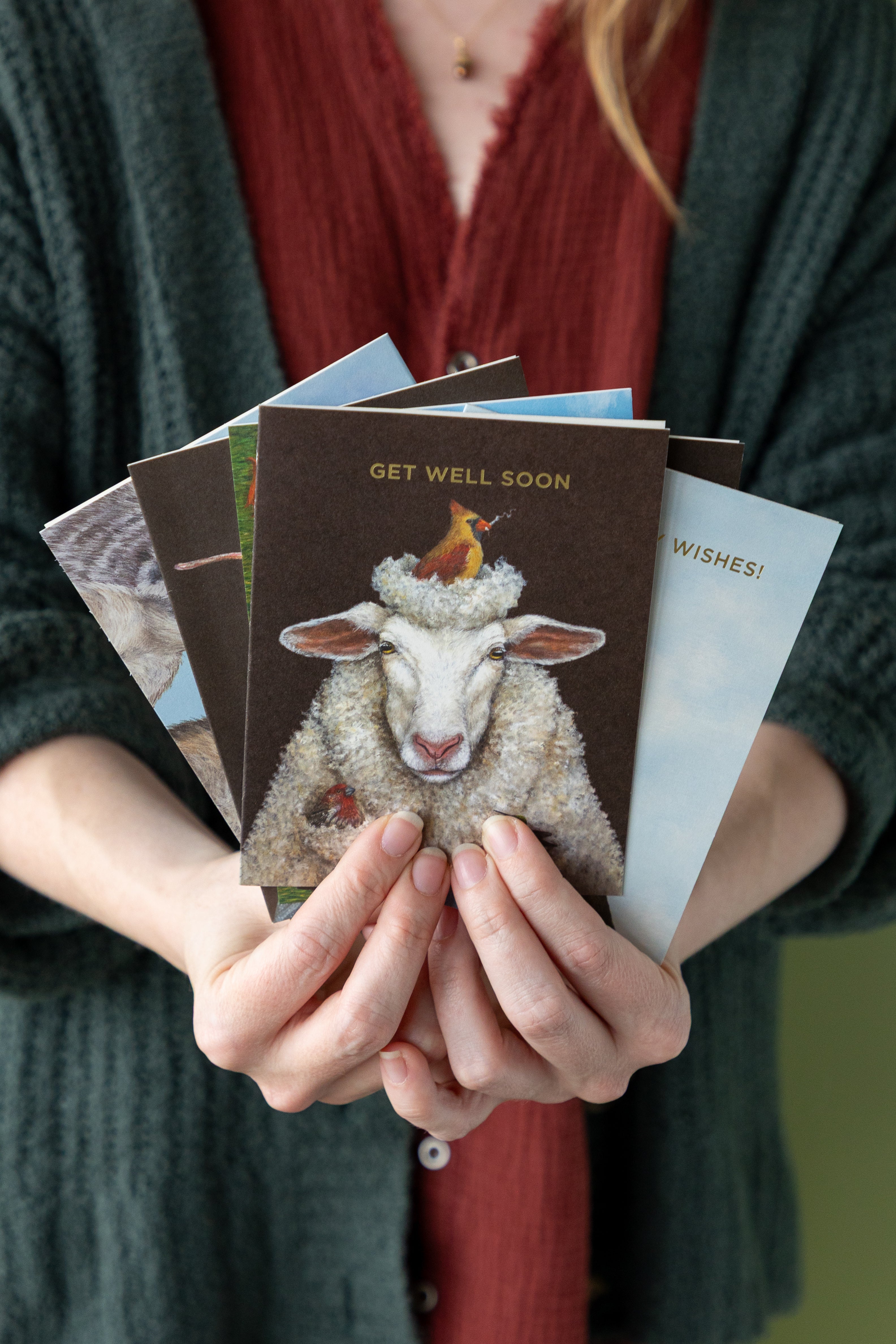 Person holding a stack of greeting cards with various designs, including one with a sheep illustration and &