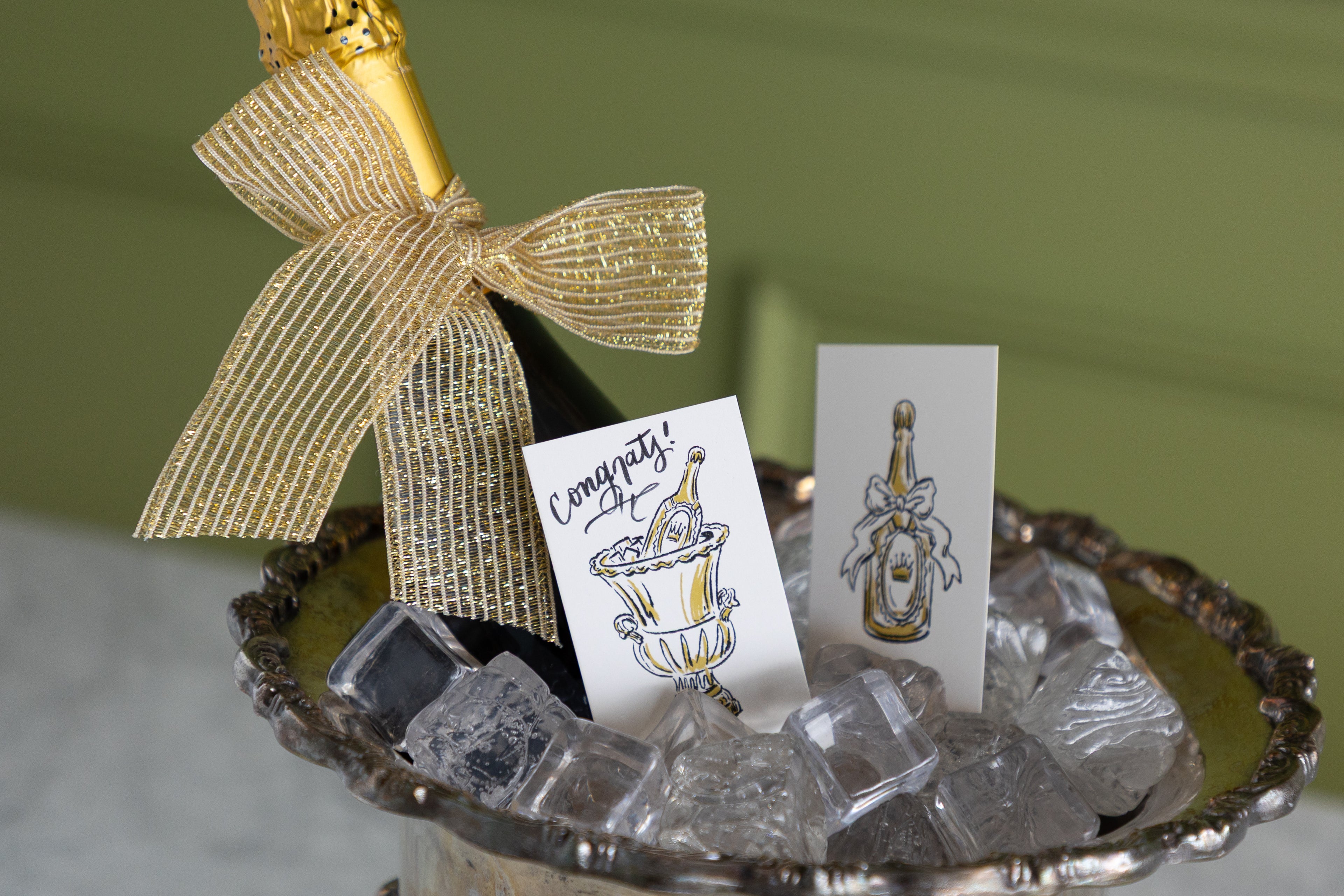 A champagne bottle with a gold foil top and a large gold ribbon bow sits in a silver tray filled with ice cubes, alongside two printed cards featuring illustrated champagne bottles and one card that says &