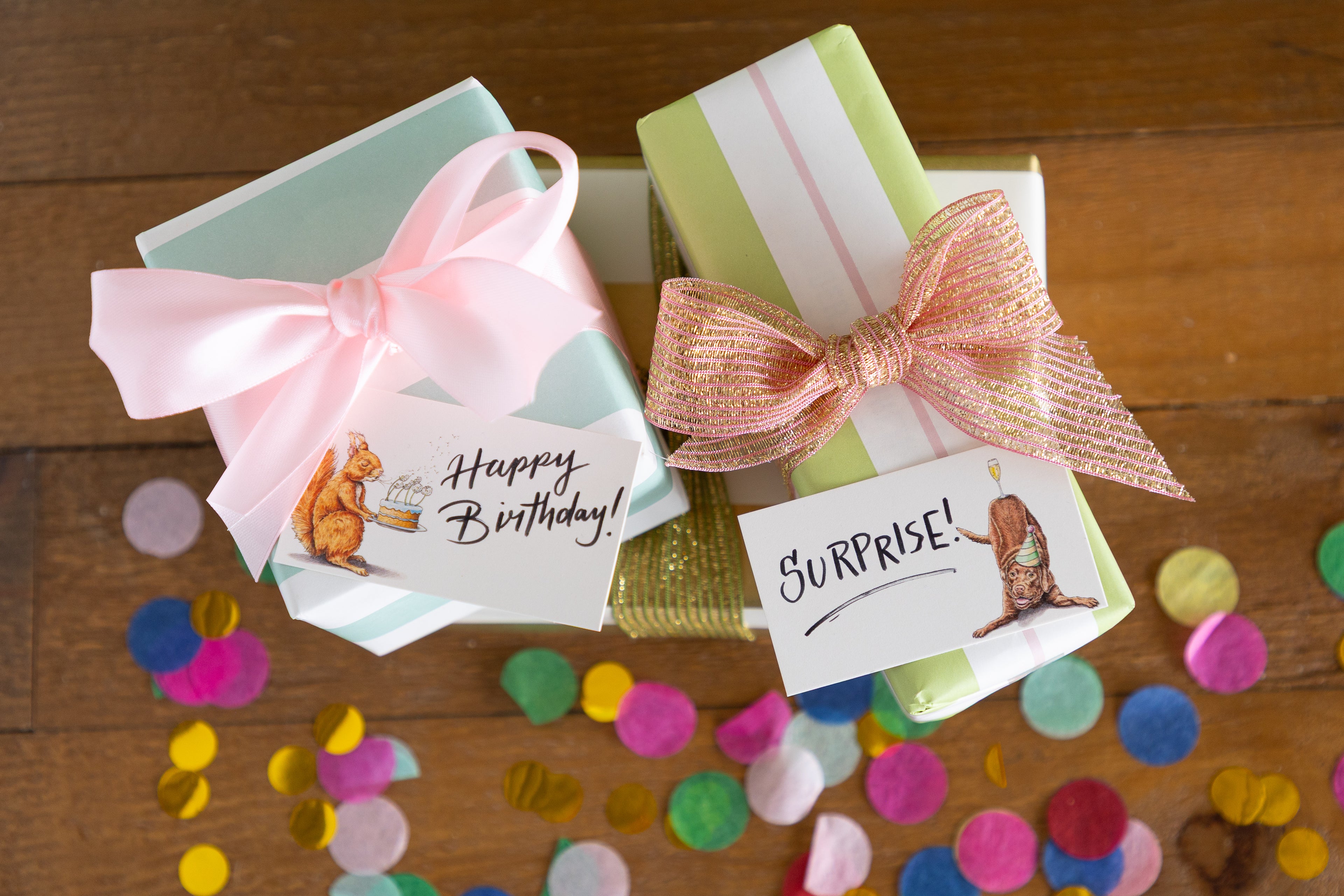 Two wrapped gift boxes on a wooden surface, one with a pale mint box, a pink ribbon, and a &