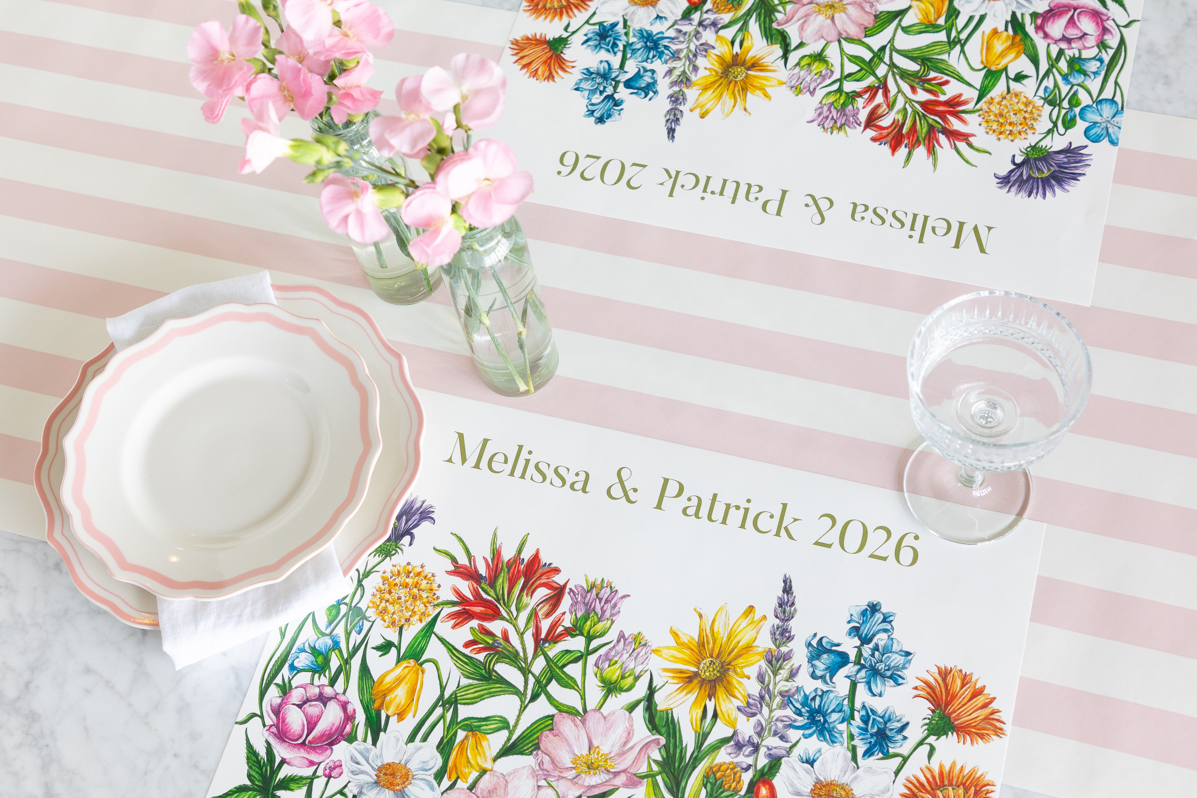 Personalized floral place mats on a pink and white striped tablecloth, styled with a white plate, a glass goblet, and pink flowers in a glass bottle.