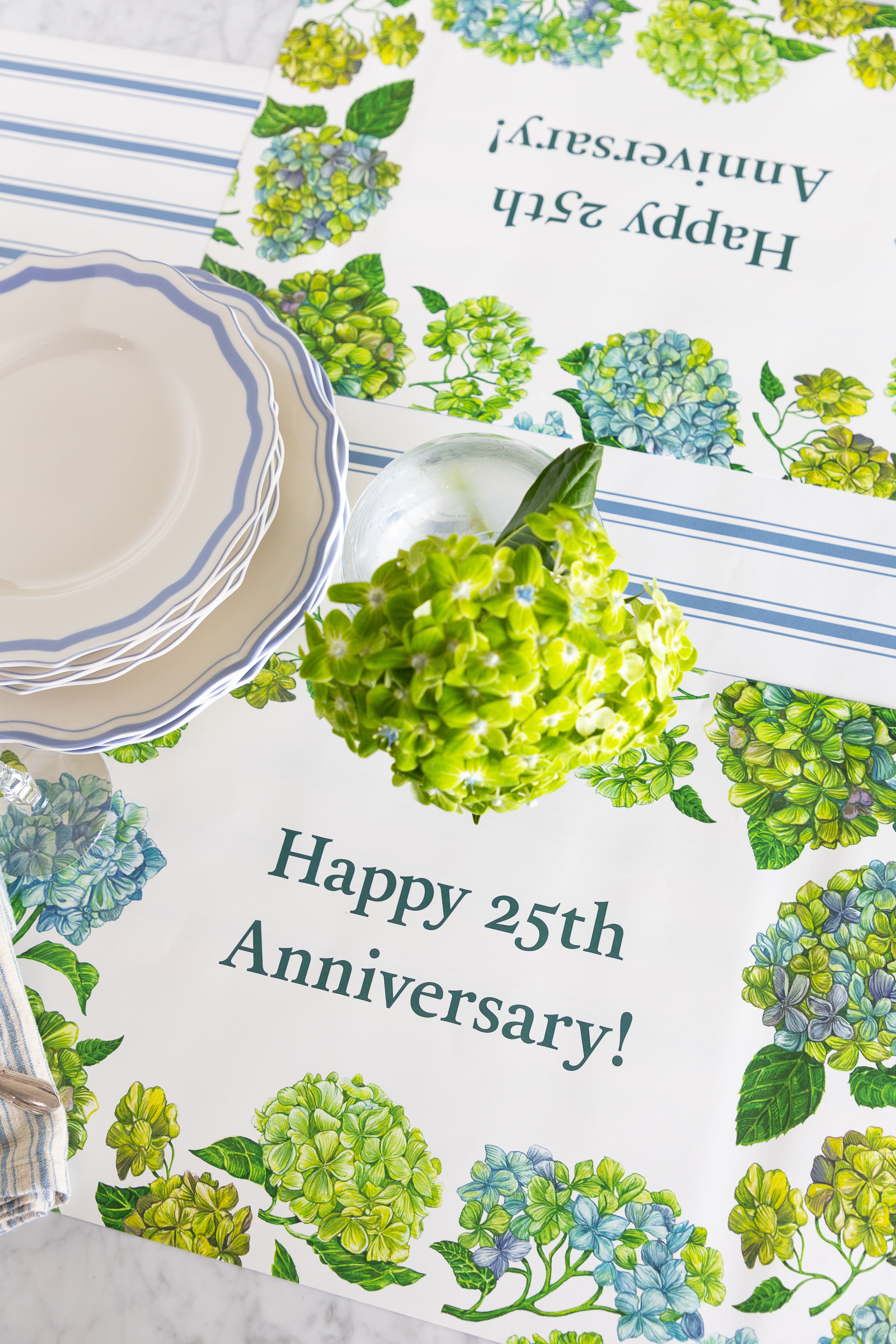 A close-up of a table setting featuring printed paper placemats with a 25th anniversary design. The placemats are decorated with watercolor-style green and blue hydrangeas and the text &