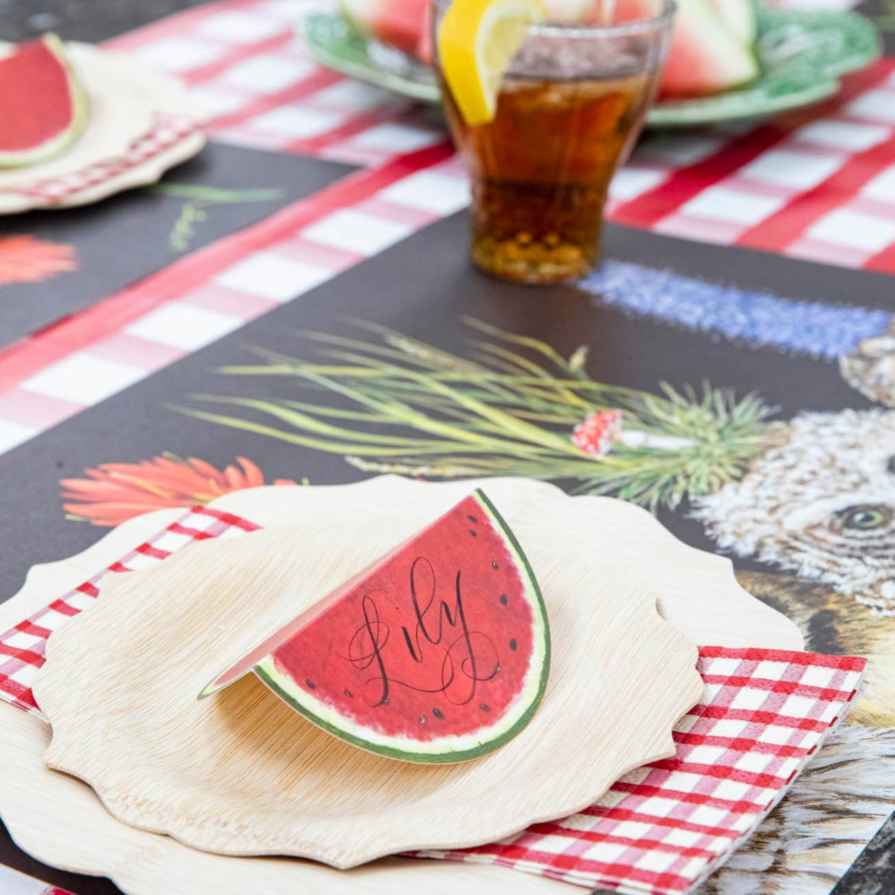 Close-up of a Watermelon Place Card labeled "Lily" standing on the plate of a summer picnic place setting.