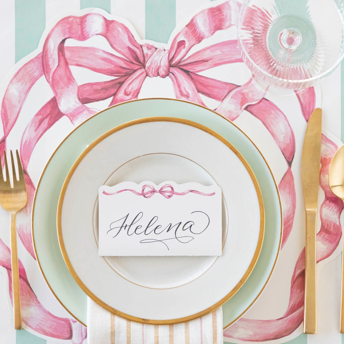 A place setting with a plate and gold cutlery, adorned with a Hester & Cook Pink Bow Place Card for added loveliness.