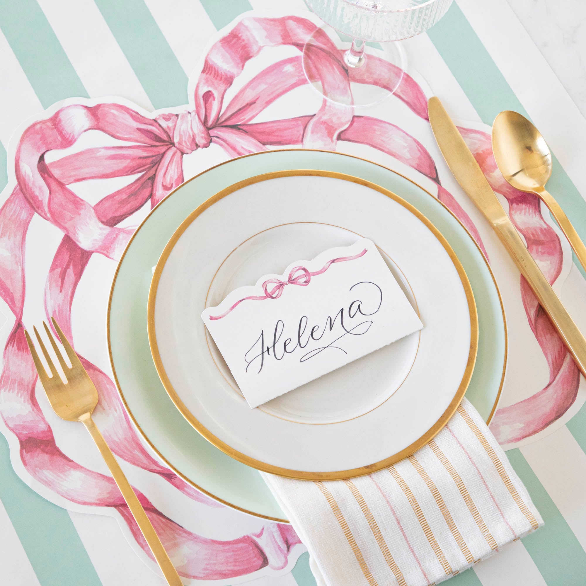 A whimsical Die-cut Pink Bow Placemat place setting with a die-cut pink bow accent, including a plate, gold spoon, and fork by Hester & Cook.