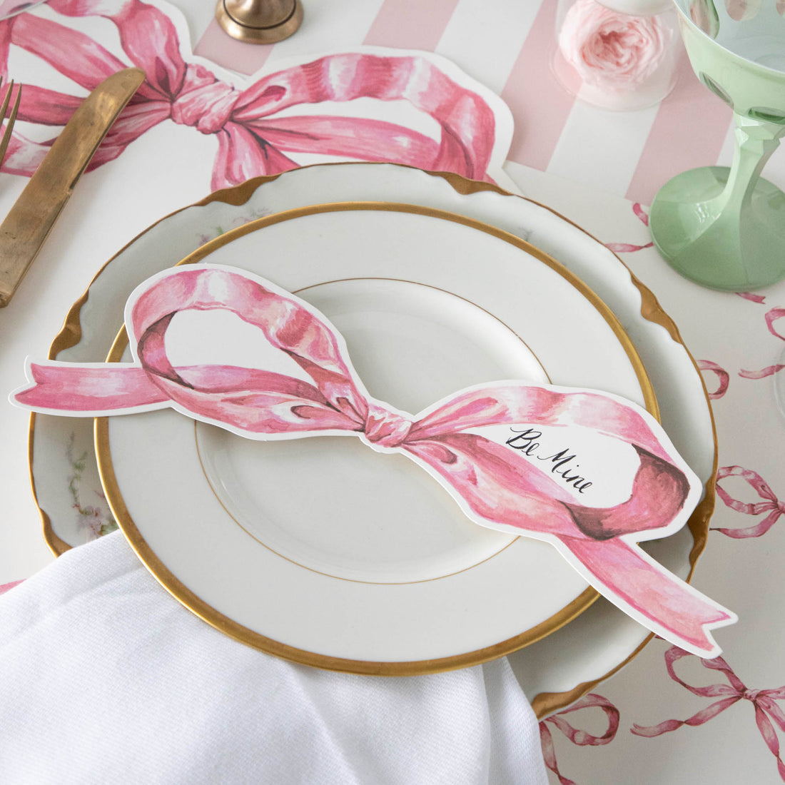 A Pink Bow Table Accent with &quot;Be Mine&quot; written in the blank space in the right bow loop resting on the plate of a pink place setting.