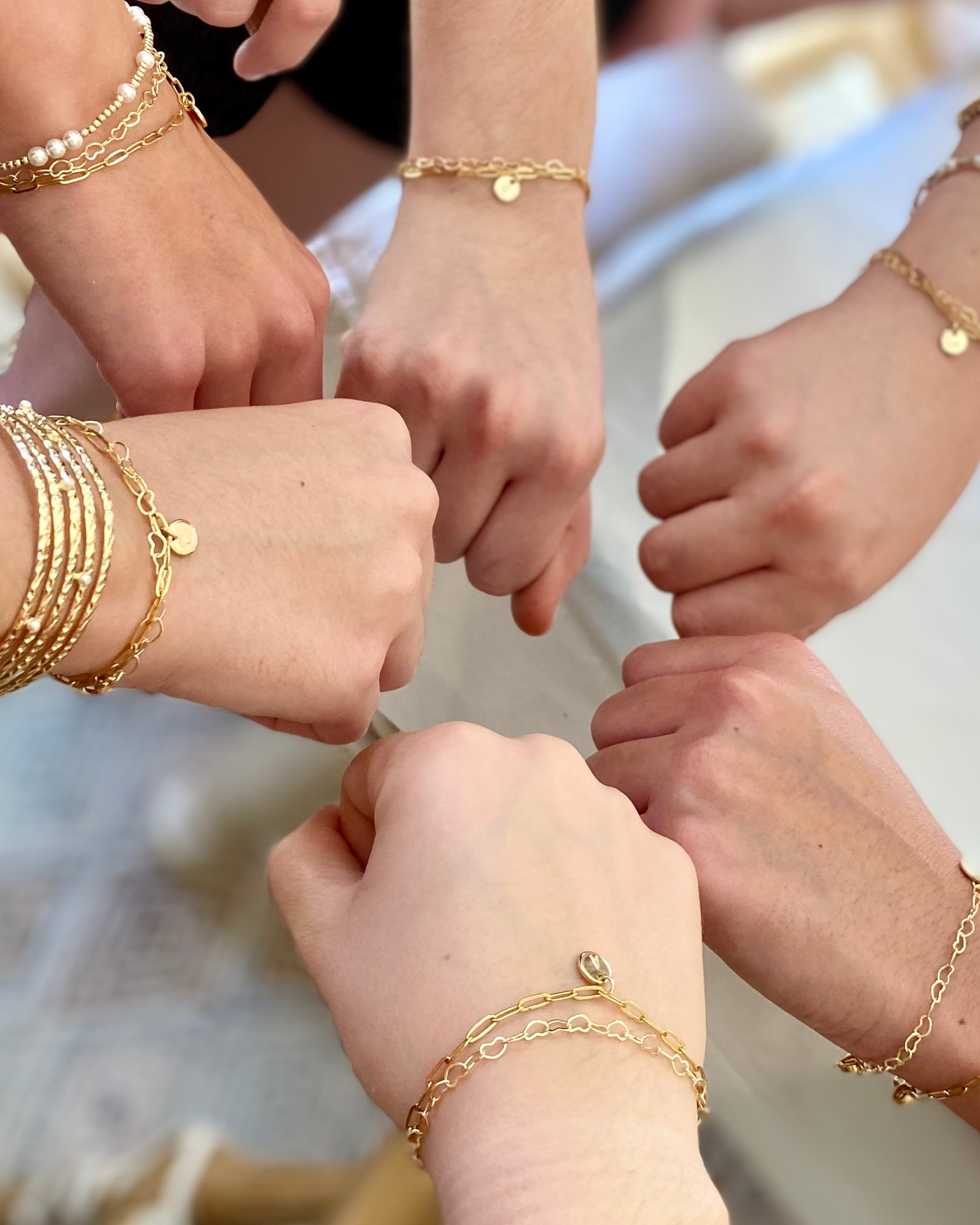 Close-up of multiple hands wearing gold bracelets on a neutral background.