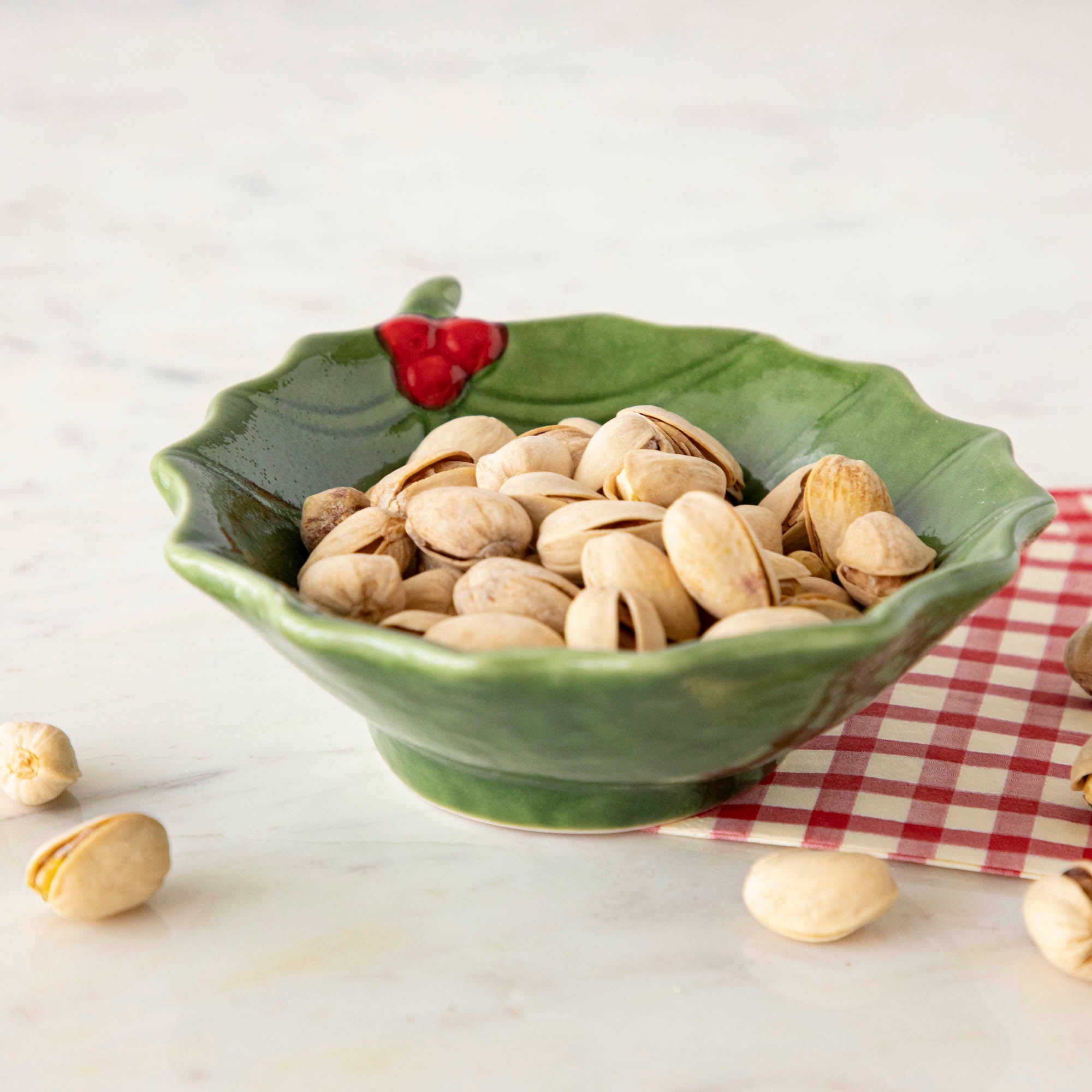 A green stoneware bowl with a holly leaf pattern filled with pistachios, placed on a red and white checkered napkin on a marble surface.
