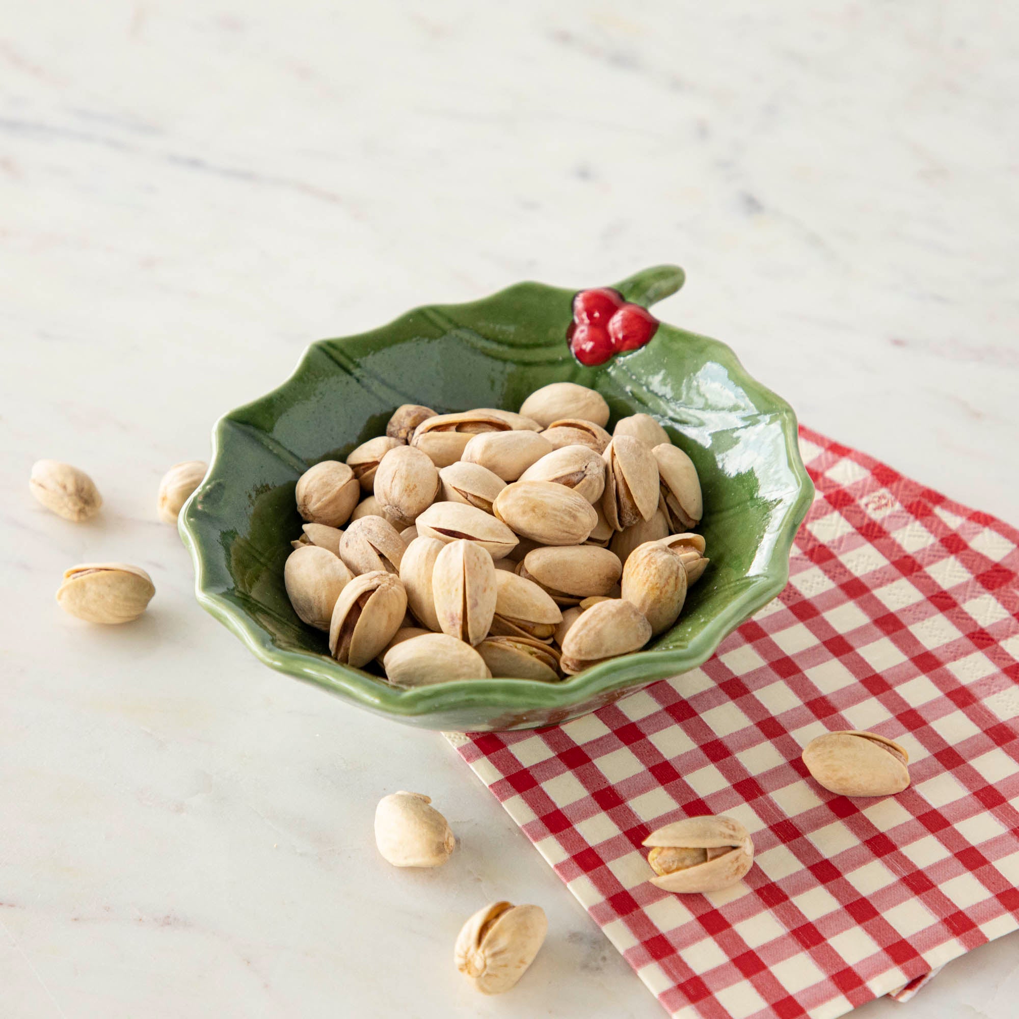 A green stoneware bowl with a holly leaf pattern filled with pistachios, placed on a red and white checkered napkin on a marble surface.