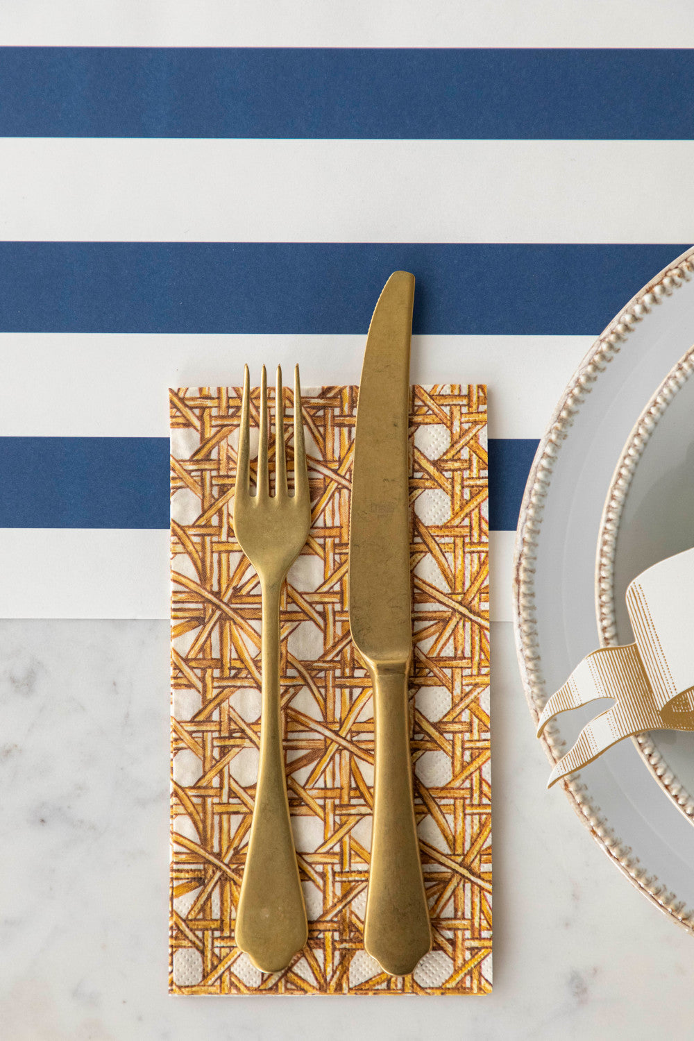Close-up of a Rattan Weave Guest Napkin under a gold cutlery set next to a plate in an elegant place setting.