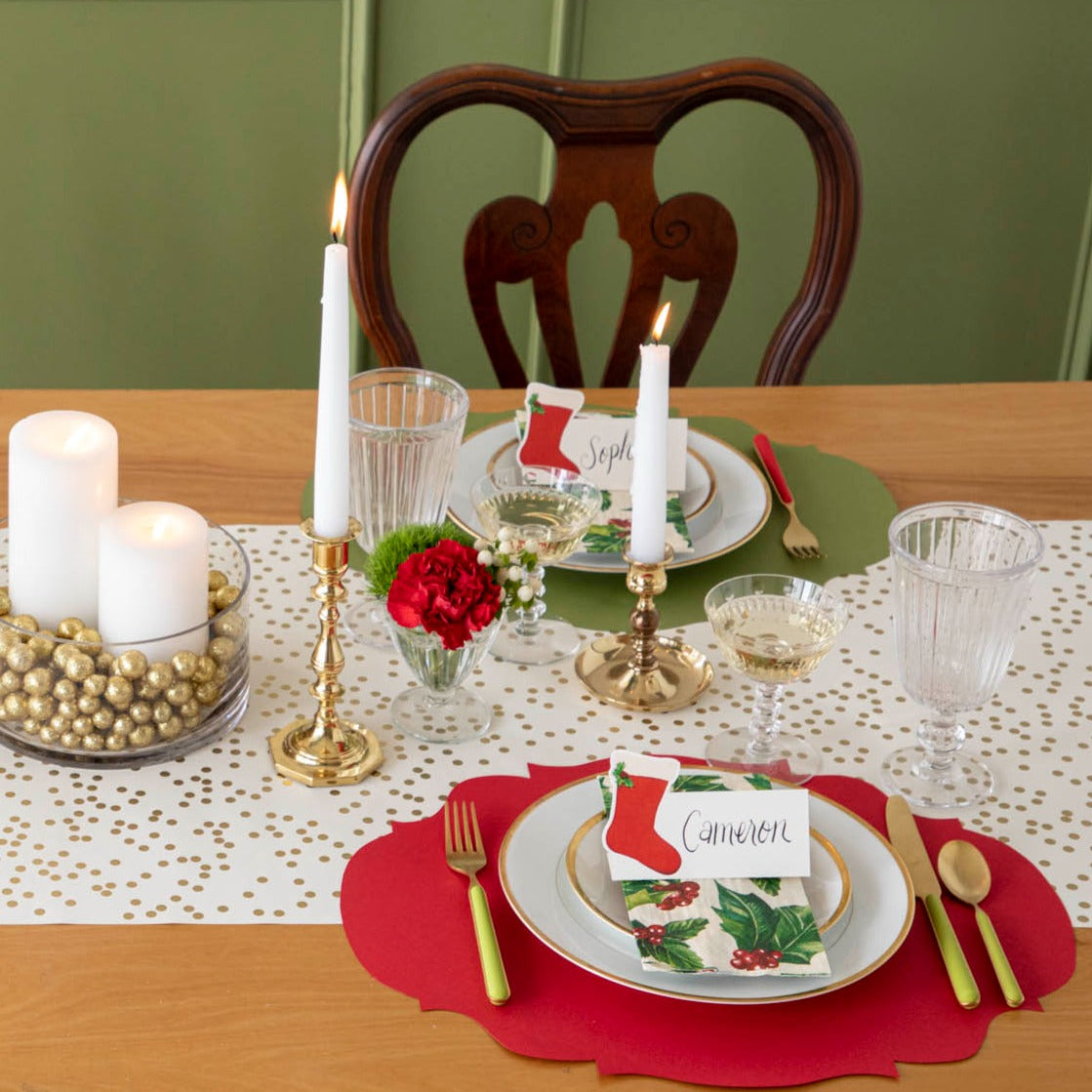 An elegant Christmas themed table setting with the Die-cut Red French Frame Placemat under gold rimmed plates, a Holly Guest Napkin, and a Stocking Place Card with the name "Cameron" written on it.