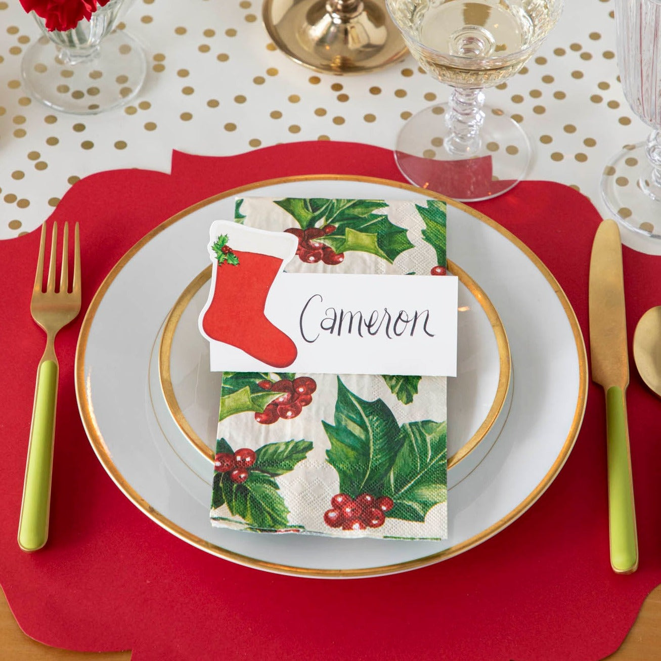 An elegant Christmas themed table setting with the Die-cut Red French Frame Placemat under gold rimmed plates, a Holly Guest Napkin, and a Stocking Place Card with the name "Cameron" written on it.