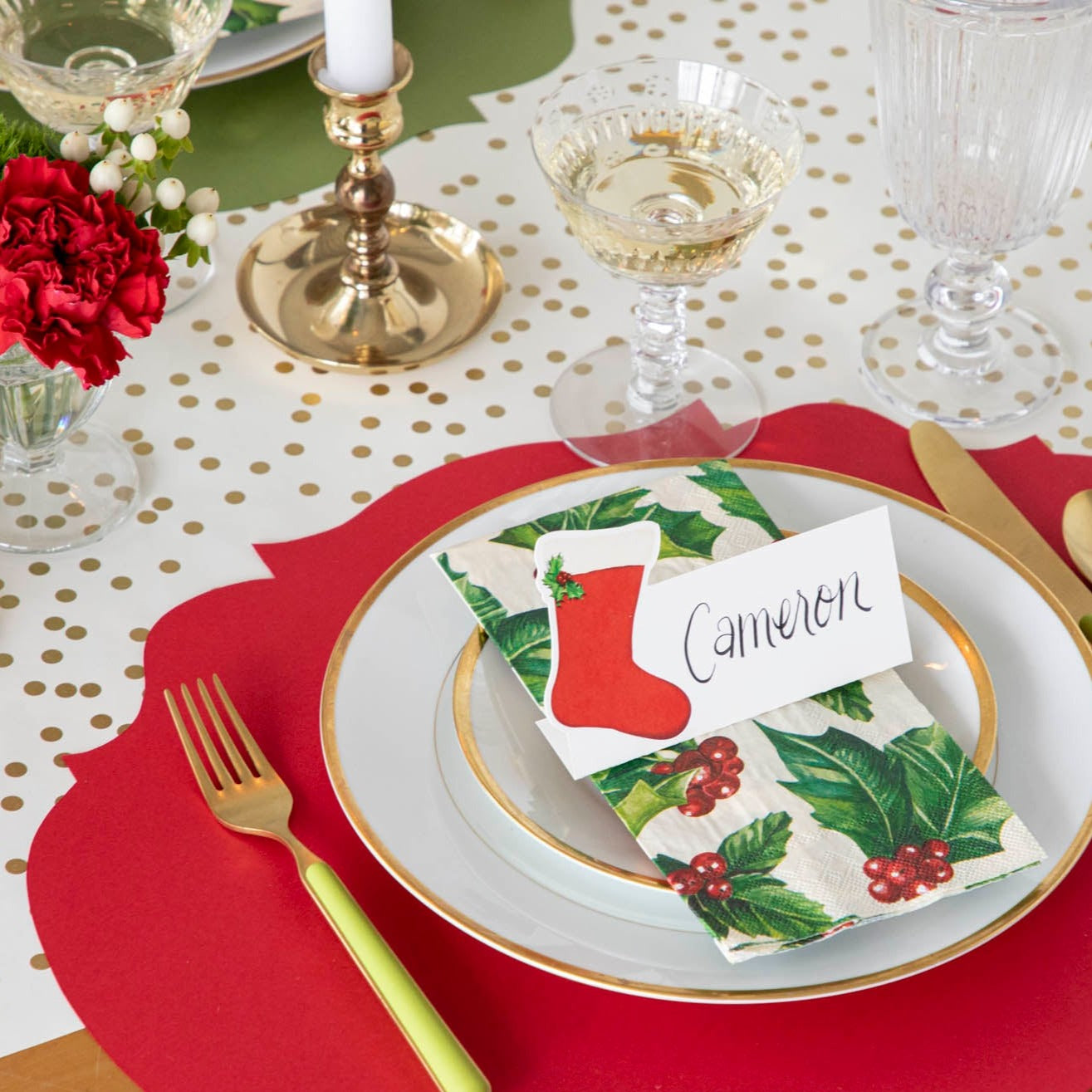 An elegant Christmas themed table setting with the Die-cut Red French Frame Placemat under gold rimmed plates, a Holly Guest Napkin, and a Stocking Place Card with the name "Cameron" written on it.