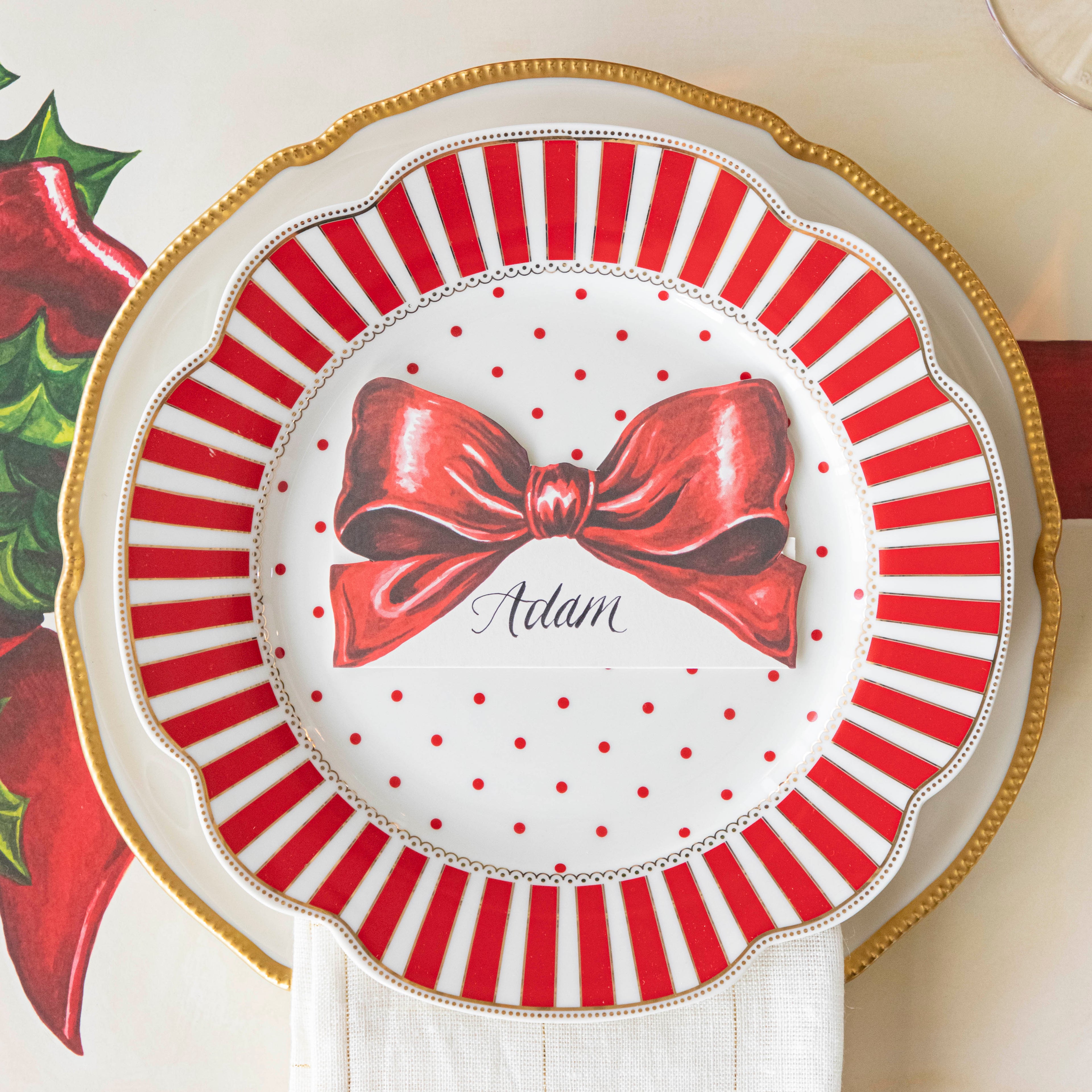 The Bow Place Card with "Adam" written on it in cursive, on a red and white striped plate with red polka dots in the center, on a gold rimmed plate with a white napkin in between.