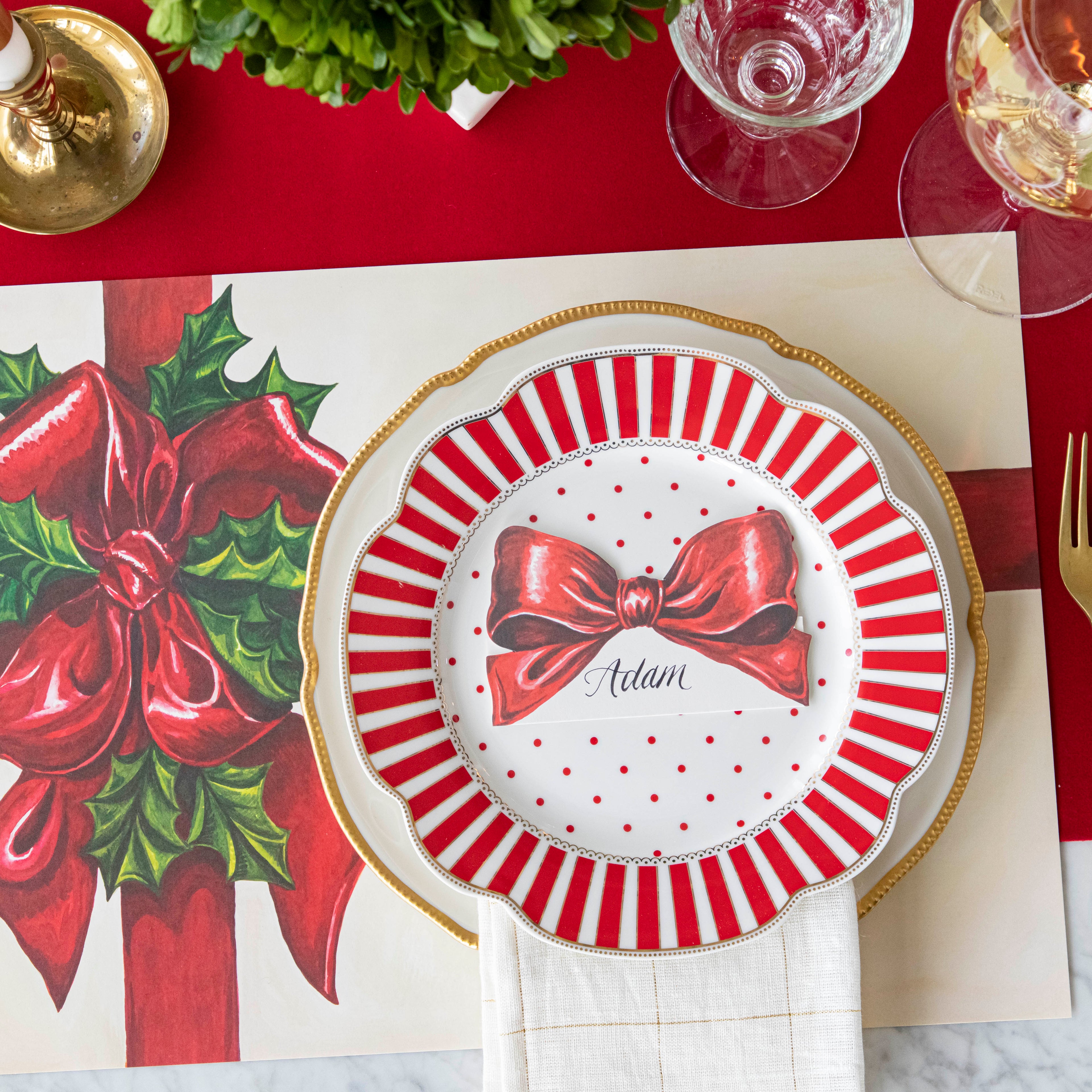 An elegant table setting with the Red Flocked Runner underneath the Christmas Present Placemat and the Bow Place Card with "Adam" written on it, resting on top of the plates.