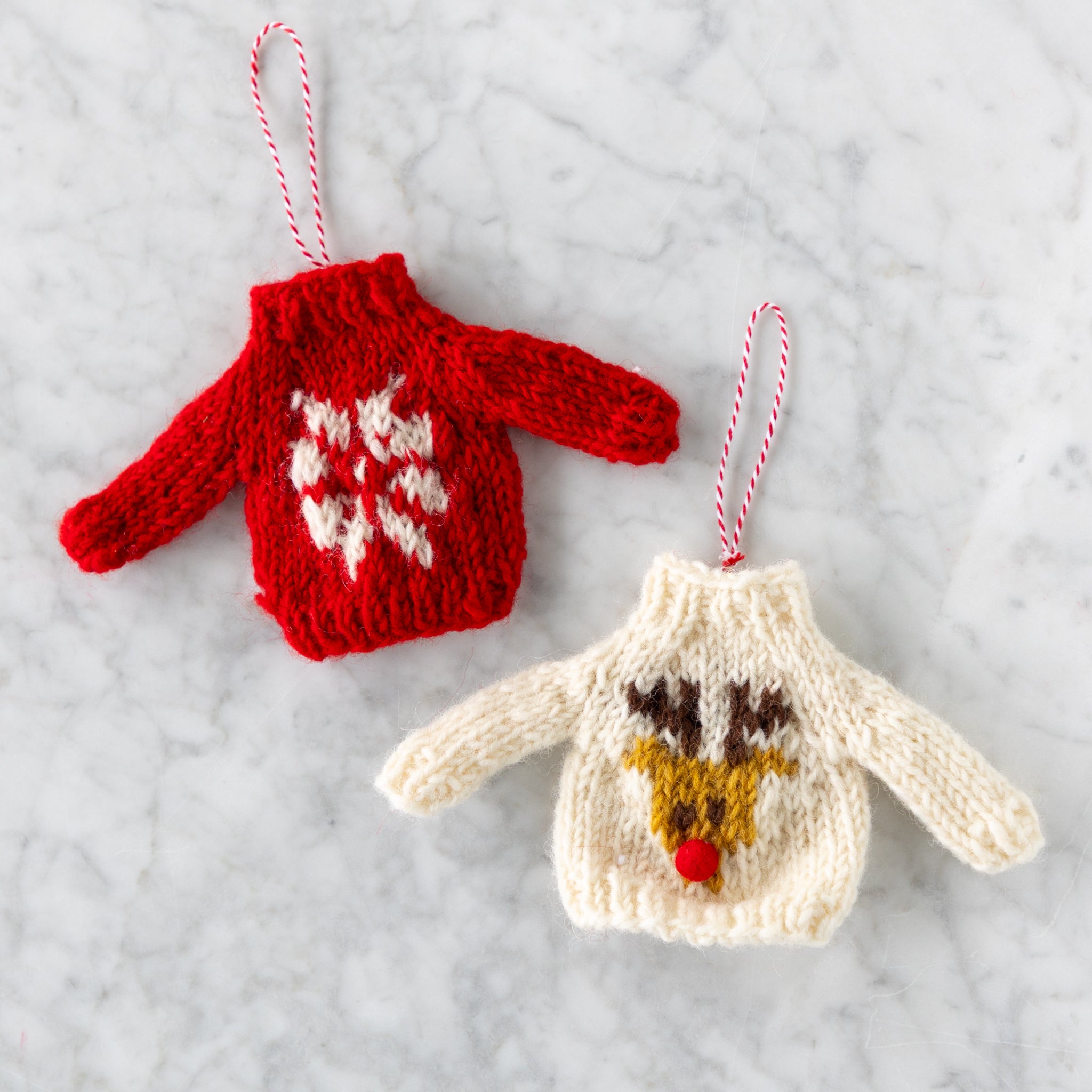 Two knitted sweater ornaments, one red with white snowflake pattern and the other cream with reindeer design, on a light gray background.