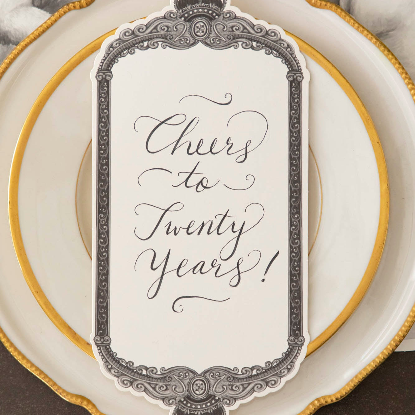 Regal Frame Table Card with "Cheers to Twenty Years!" written on it, resting on dinner plates.