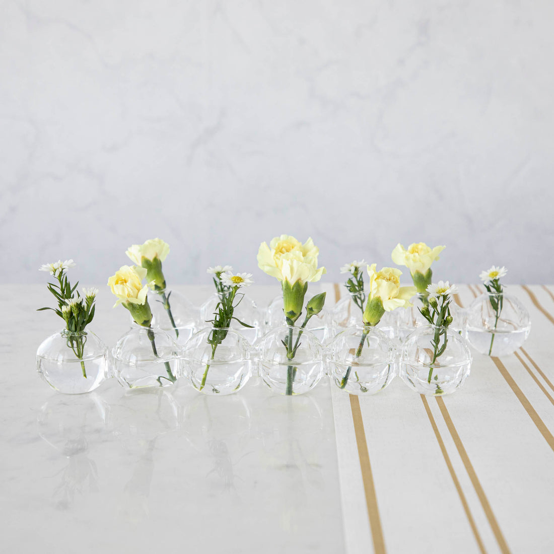 A row of small Chive Caterpillar Vases each containing a single yellow carnation, arranged on a striped surface against a marble background.