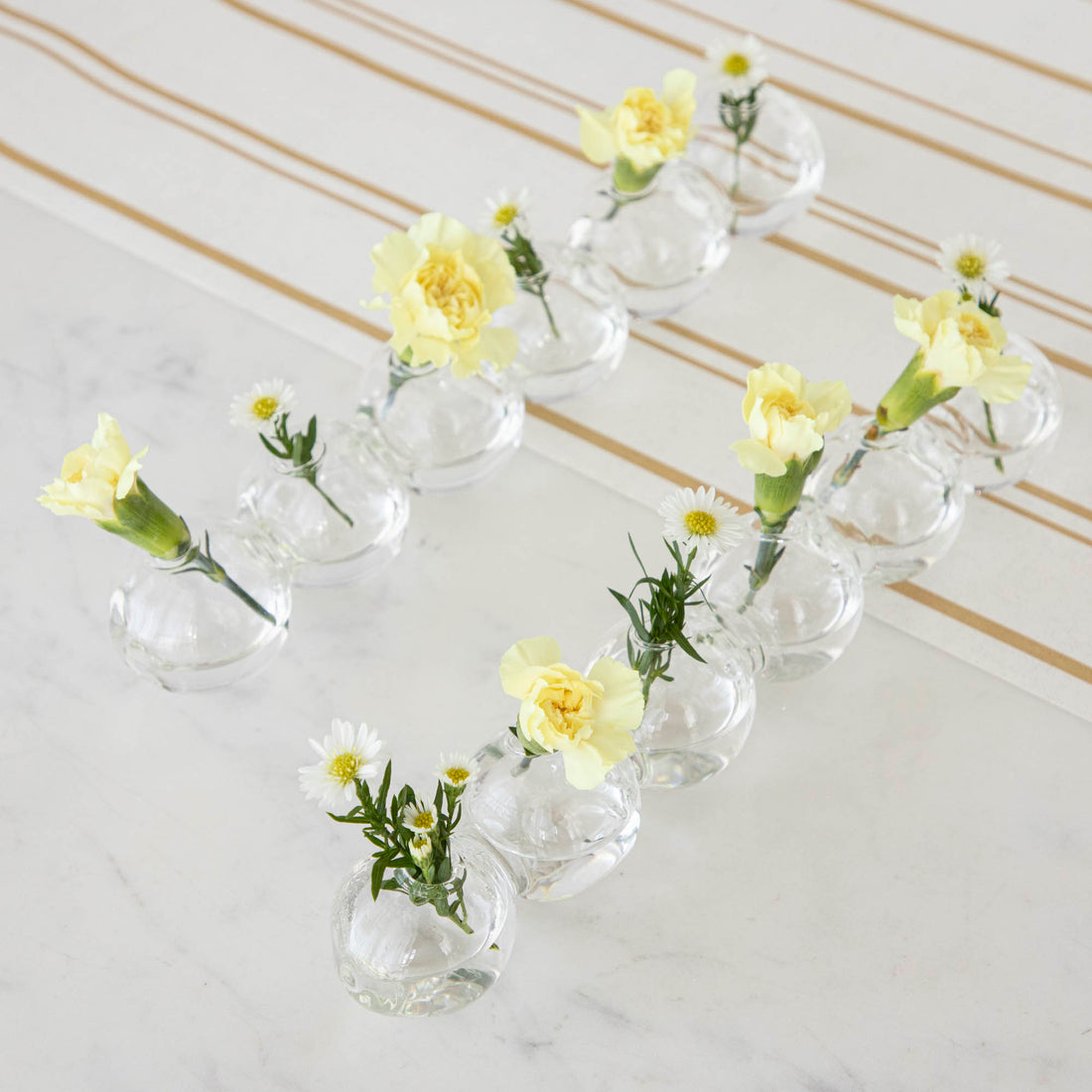 A row of small Chive Caterpillar Vases each containing a single yellow carnation, arranged on a striped surface against a marble background.