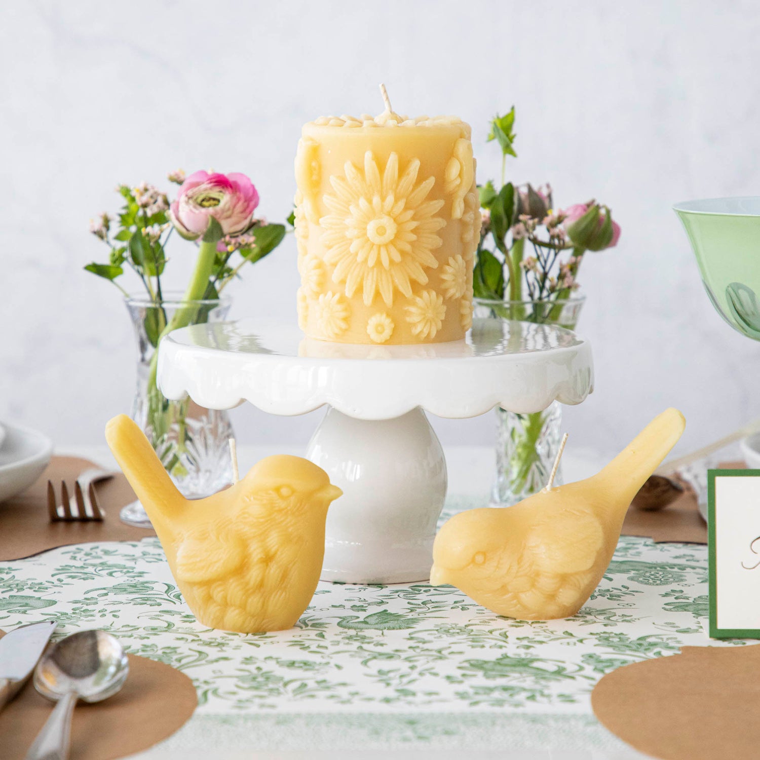 A Beeswax Floral Pillar Candle resting on a cake stand on a table with two yellow bird candles with pink flowers in the background.