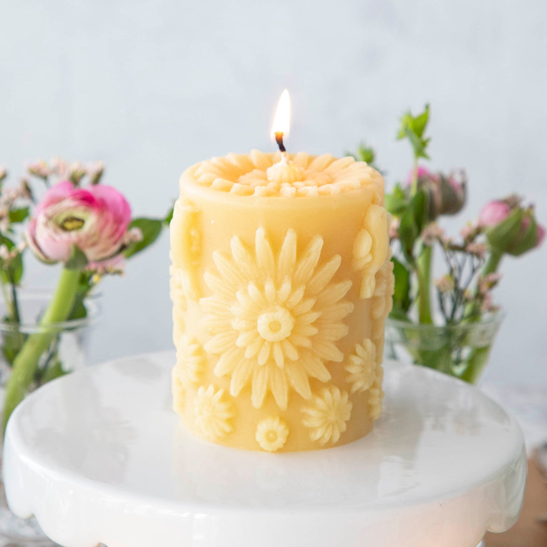 A lit, yellow beeswax pillar candle covered with large and small 3D circular flowers, including a flower on the top surface, resting on a cake stand with pink flowers in the background.
