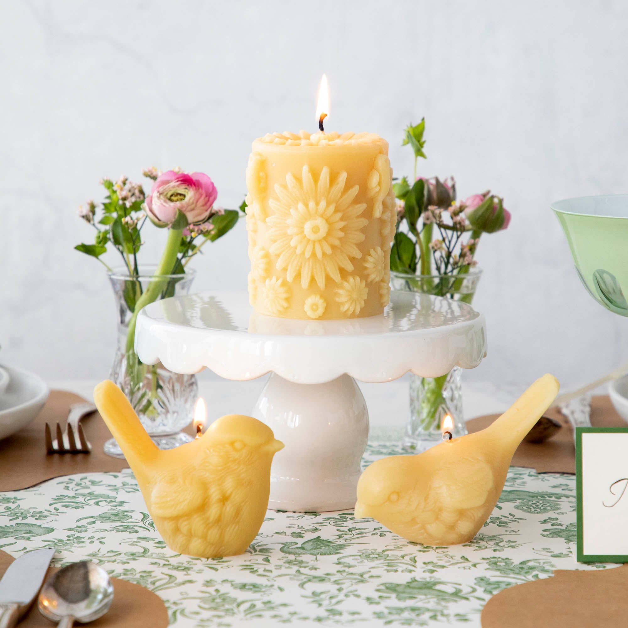 A Beeswax Floral Pillar Candle resting on a cake stand on a table with two yellow bird candles with pink flowers in the background.