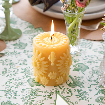A lit, yellow beeswax pillar candle covered with large and small 3D circular flowers, including a flower on the top surface, resting on a table setting with flowers in vases.