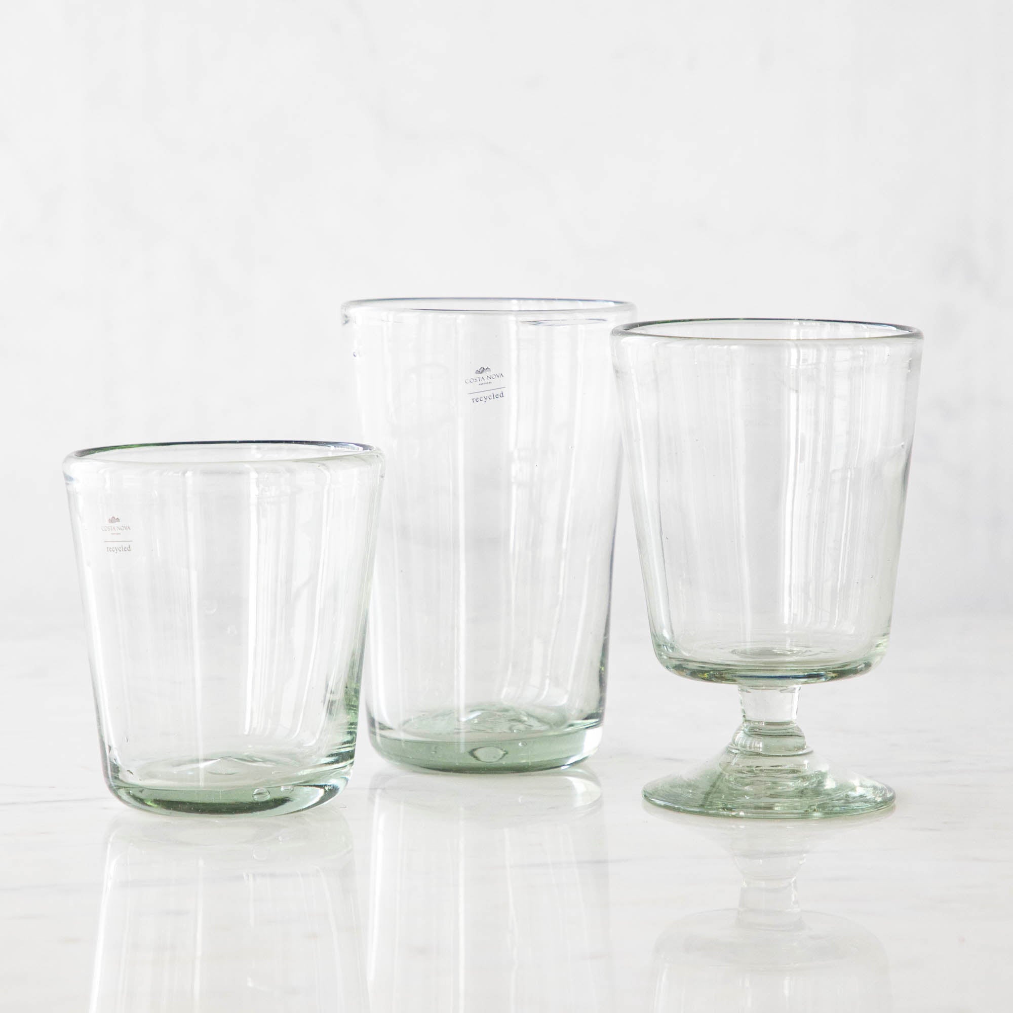 Three empty Margarida Recycled Glasses vessels of varying heights and shapes, made by Casafina Living, rest on a white surface.