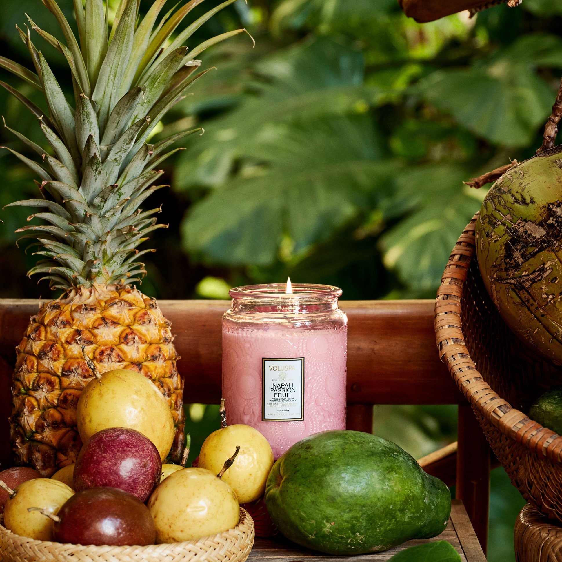 Pink candle with a label on a wooden surface surrounded by fruits and a pineapple, with a blurred green leafy background.