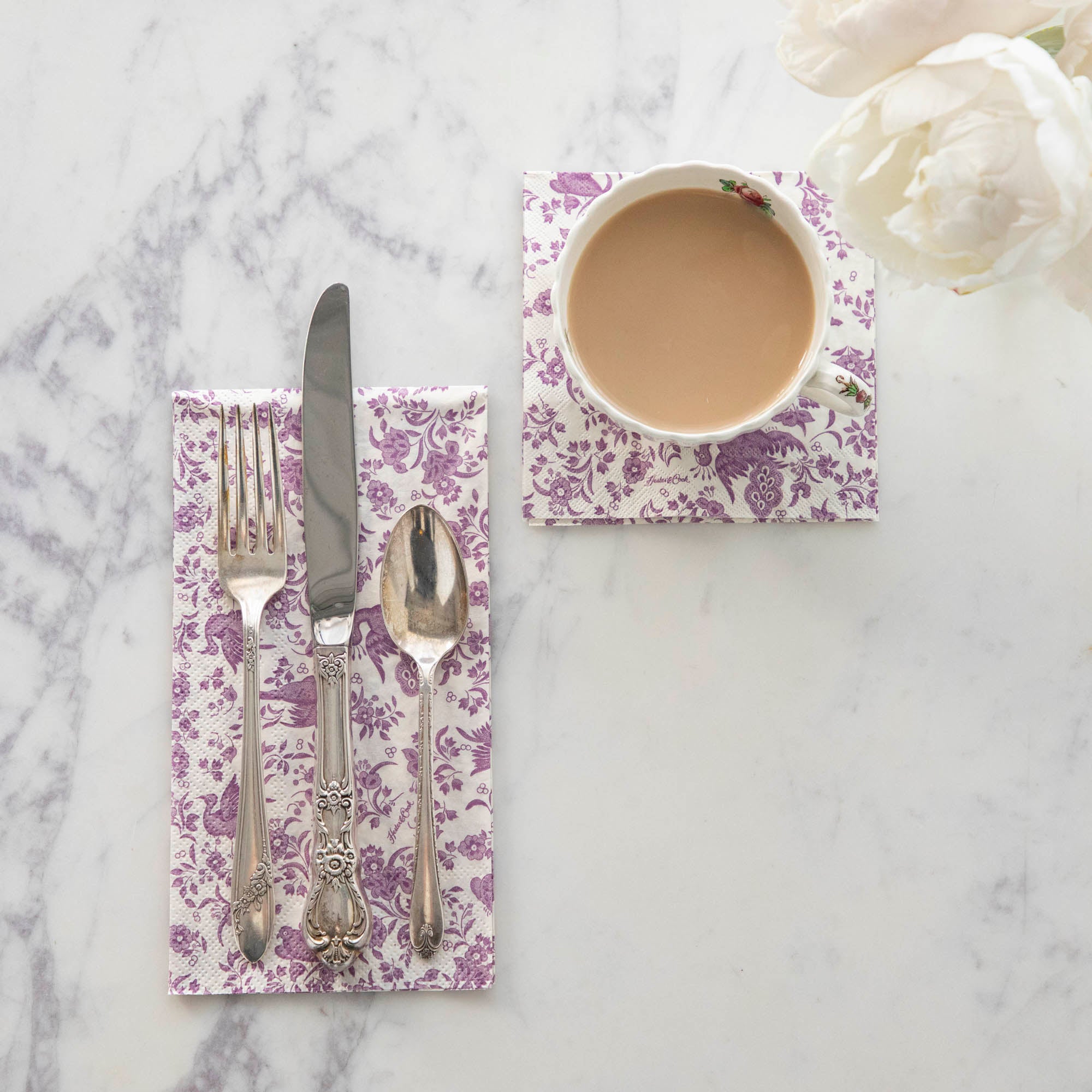 A top-down view of a set of cutlery on a Lilac Regal Peacock Guest Napkin next to a cup of coffee on a Lilac Regal Peacock Cocktail Napkin.