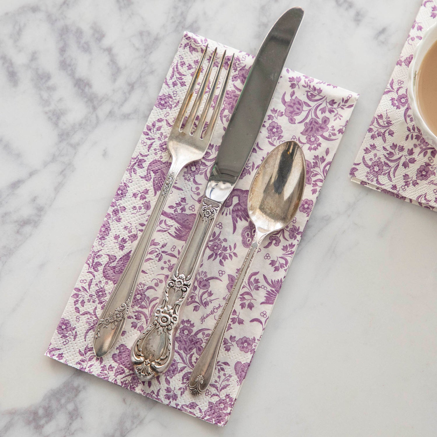 A Lilac Regal Peacock Guest Napkin by Hester &amp; Cook with a set of cutlery on it.