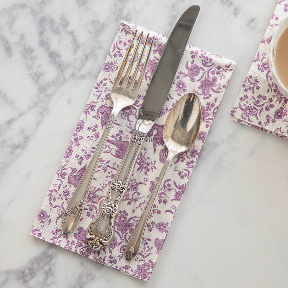 A Lilac Regal Peacock Guest Napkin by Hester & Cook with a set of cutlery on it.