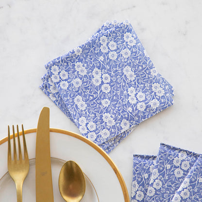 A stack of three Blue Calico Cocktail Napkins fanned out next to a gold-rimmed plate with gold flatware.